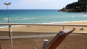 A beach view from a balcony with a chair and a table, a railing, and a distant view of the sea and a rocky hill.