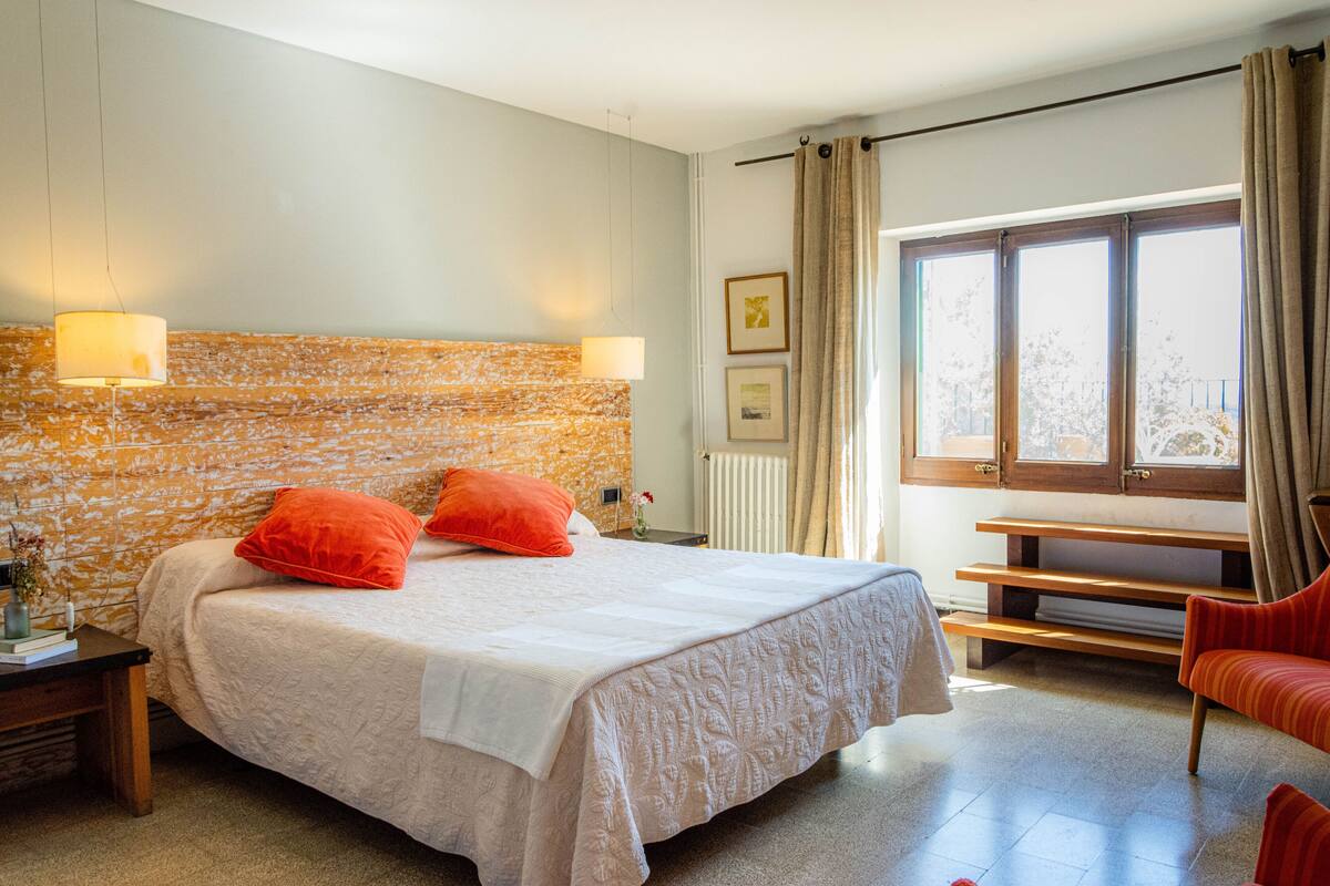 A bedroom with a large bed, two bedside lamps, a window with curtains, a bench, and a chair.