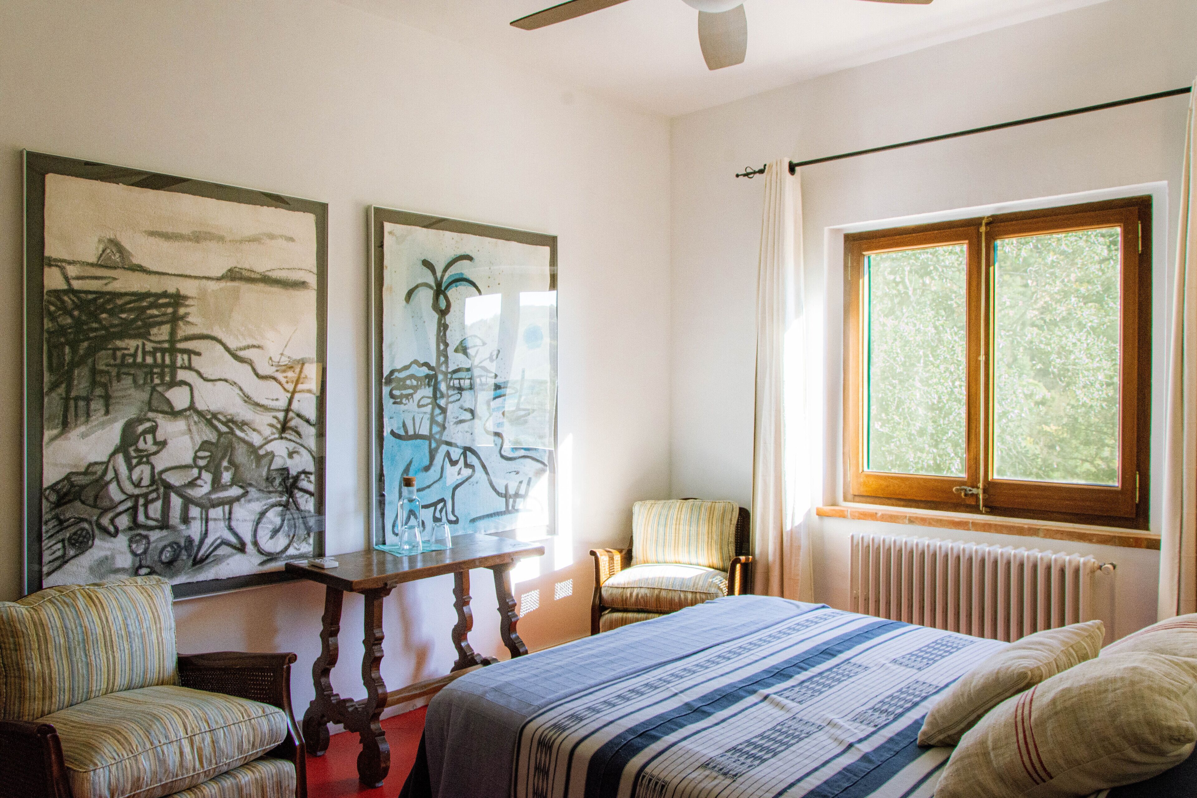 A room with a bed, two chairs, a table, and two paintings on the wall.