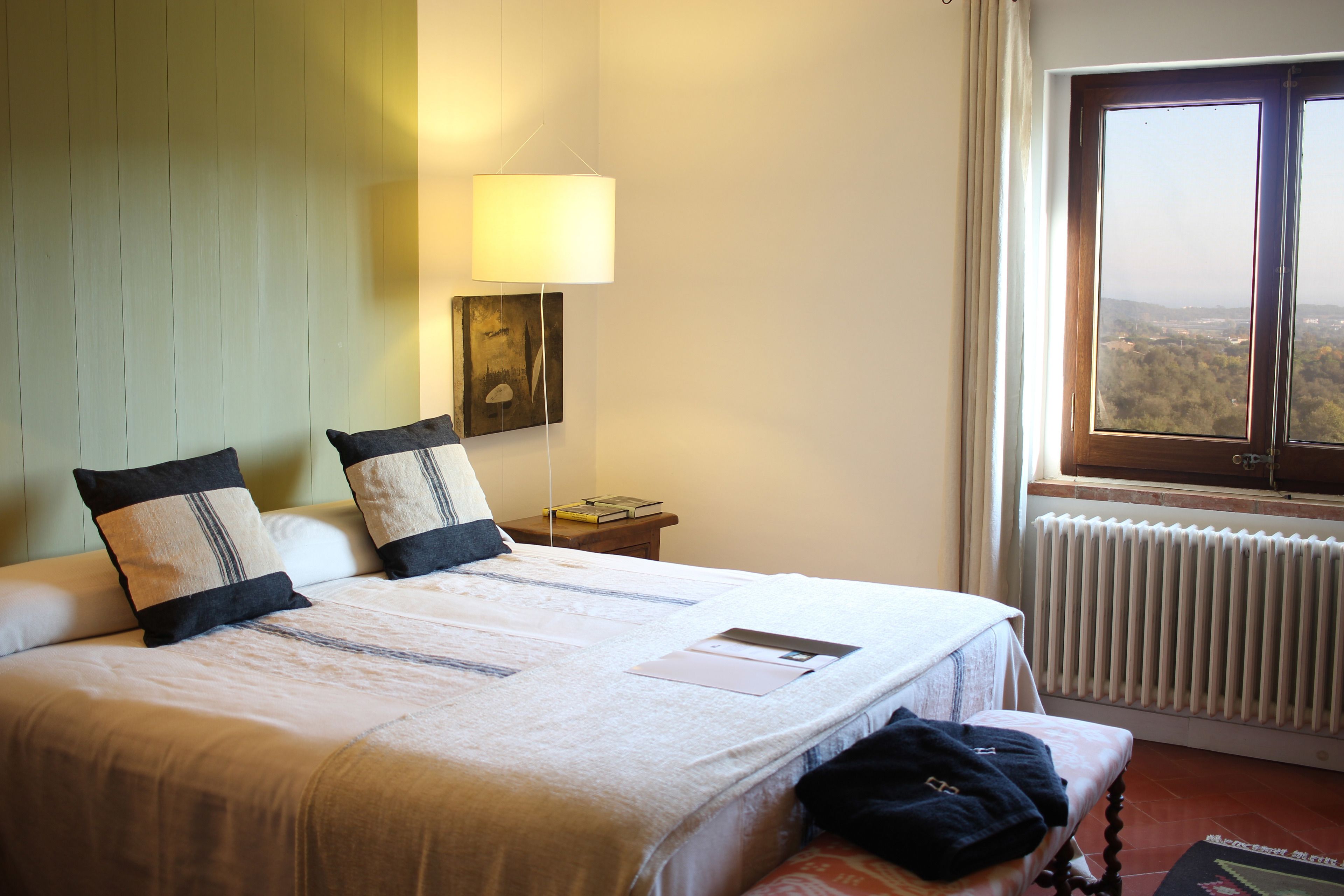 A neatly made bed with a white and beige bedspread, two pillows, a bedside table with a lamp, and a view of a cityscape through a window.