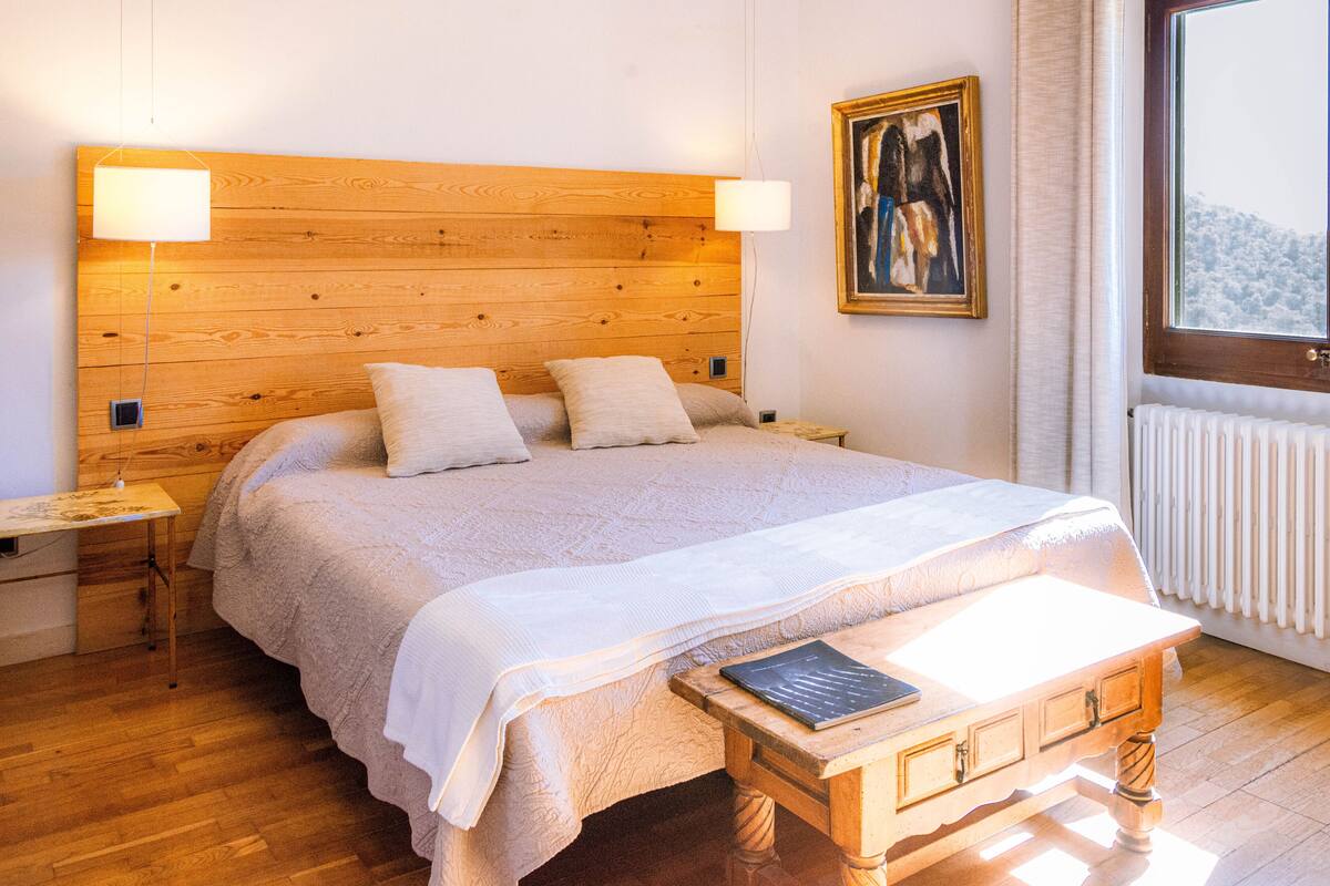 A bedroom with a wooden headboard, a large bed with a white bedspread, two bedside tables, a nightstand, a bedside lamp, a framed picture on the wall, and a window with a view of a snowy landscape.