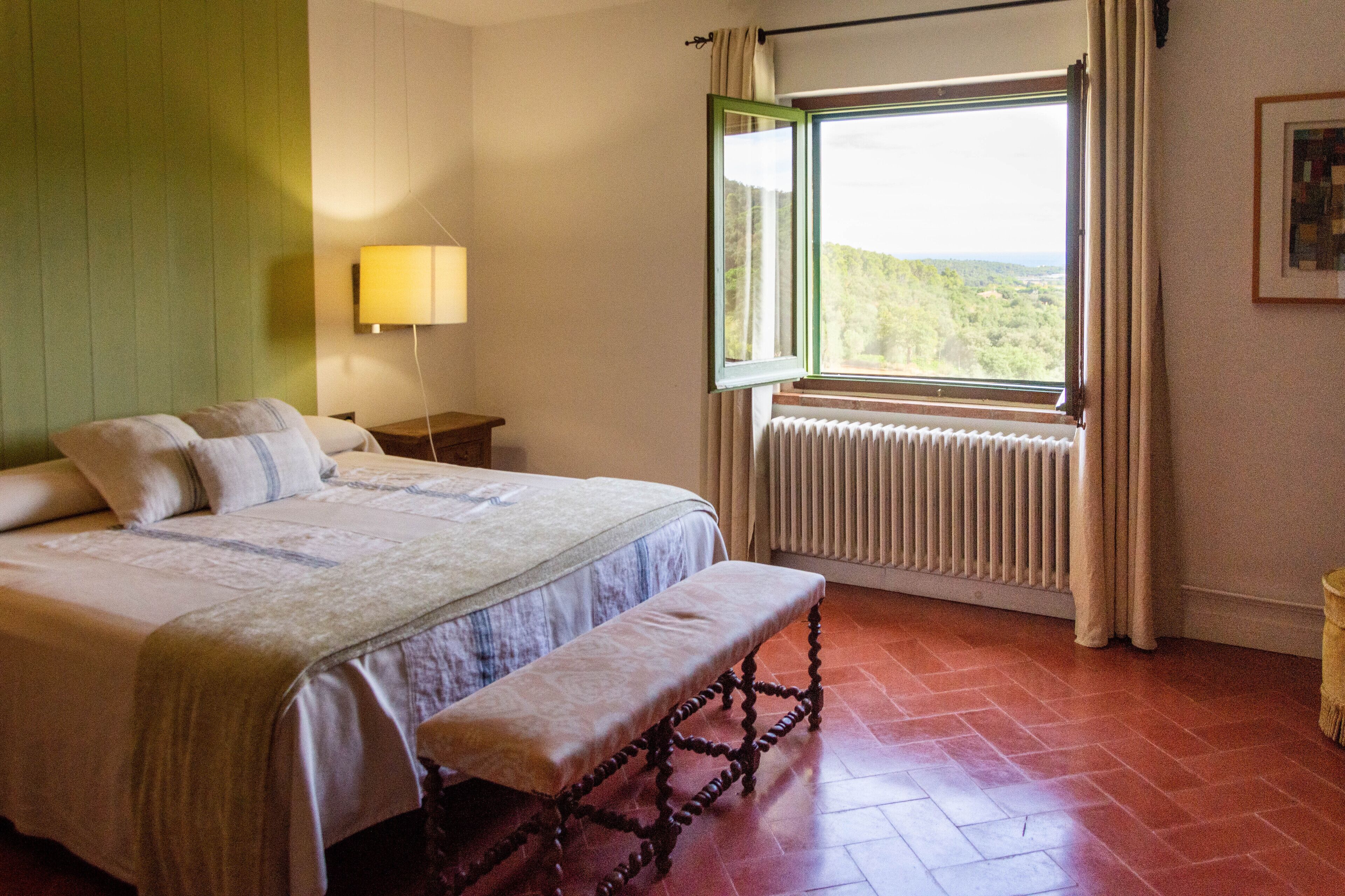 Une chambre à coucher comprenant un lit, un banc, une table de chevet, une lampe, une fenêtre, un radiateur et une vue sur la verdure à l’extérieur.