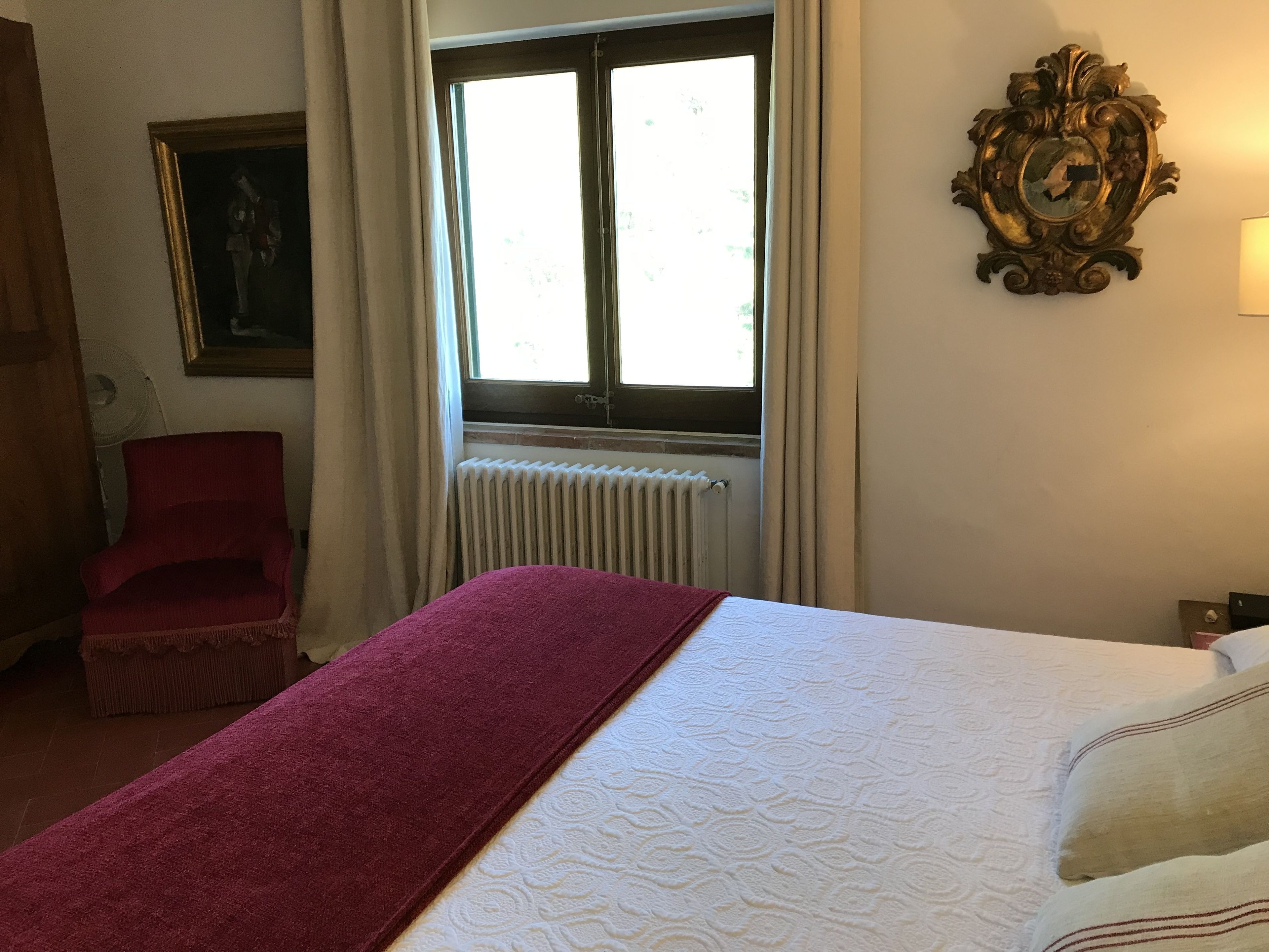 A hotel room with a bed, a red chair, a window with curtains, a radiator, and a wall-mounted clock.