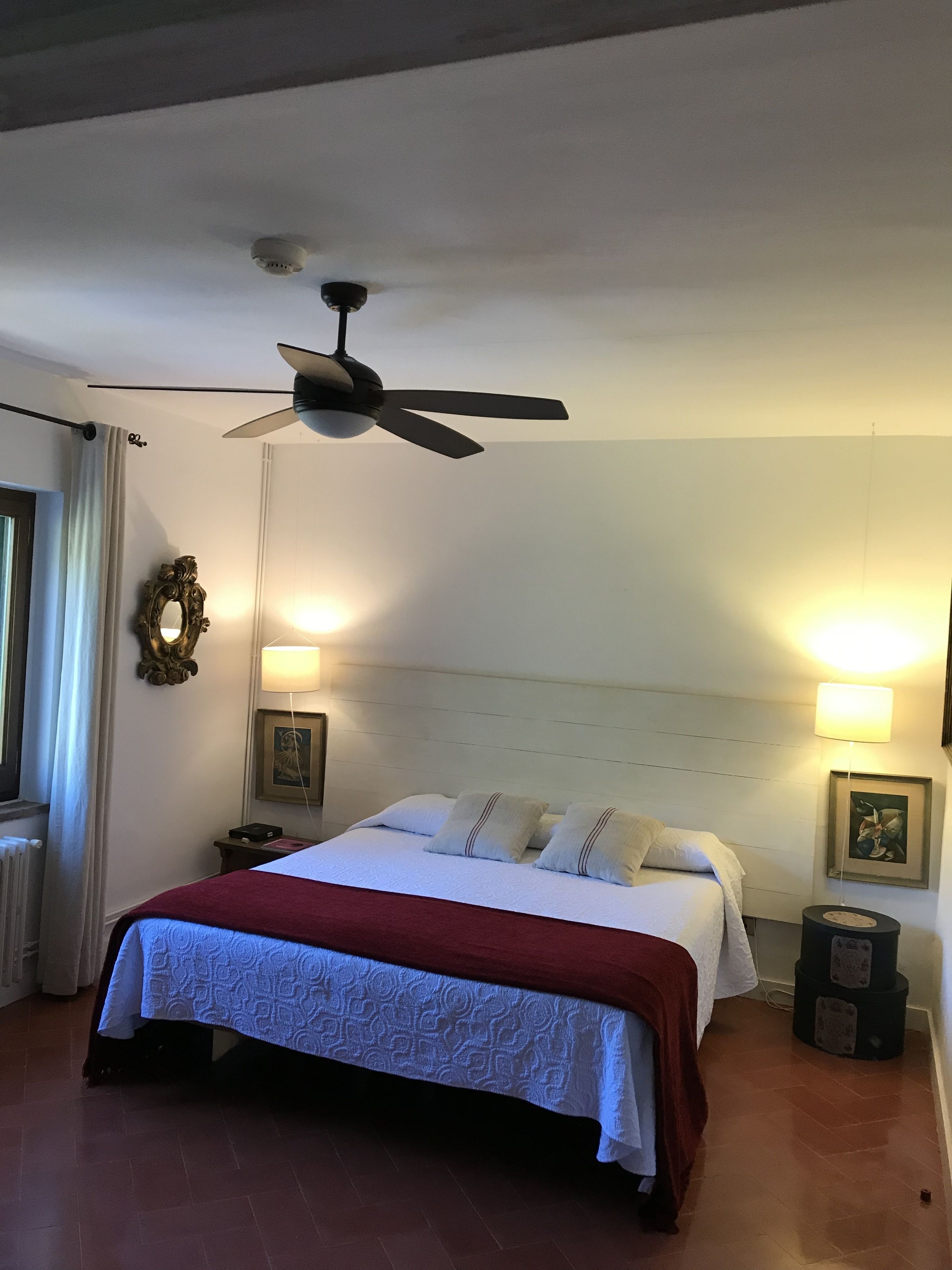 Habitación de hotel con cama, ventilador de techo, lámparas de noche y un cuadro en la pared.