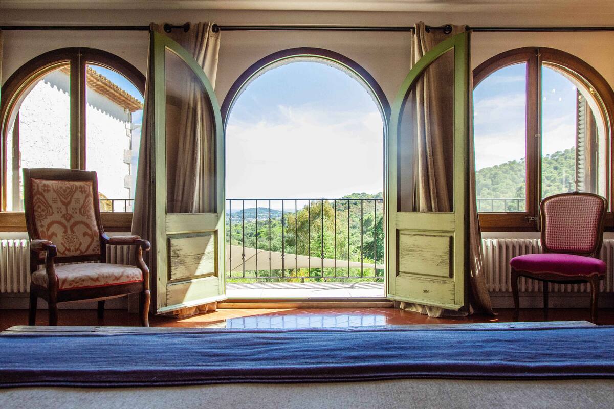 A room with a balcony, two chairs, and a view of a green landscape.