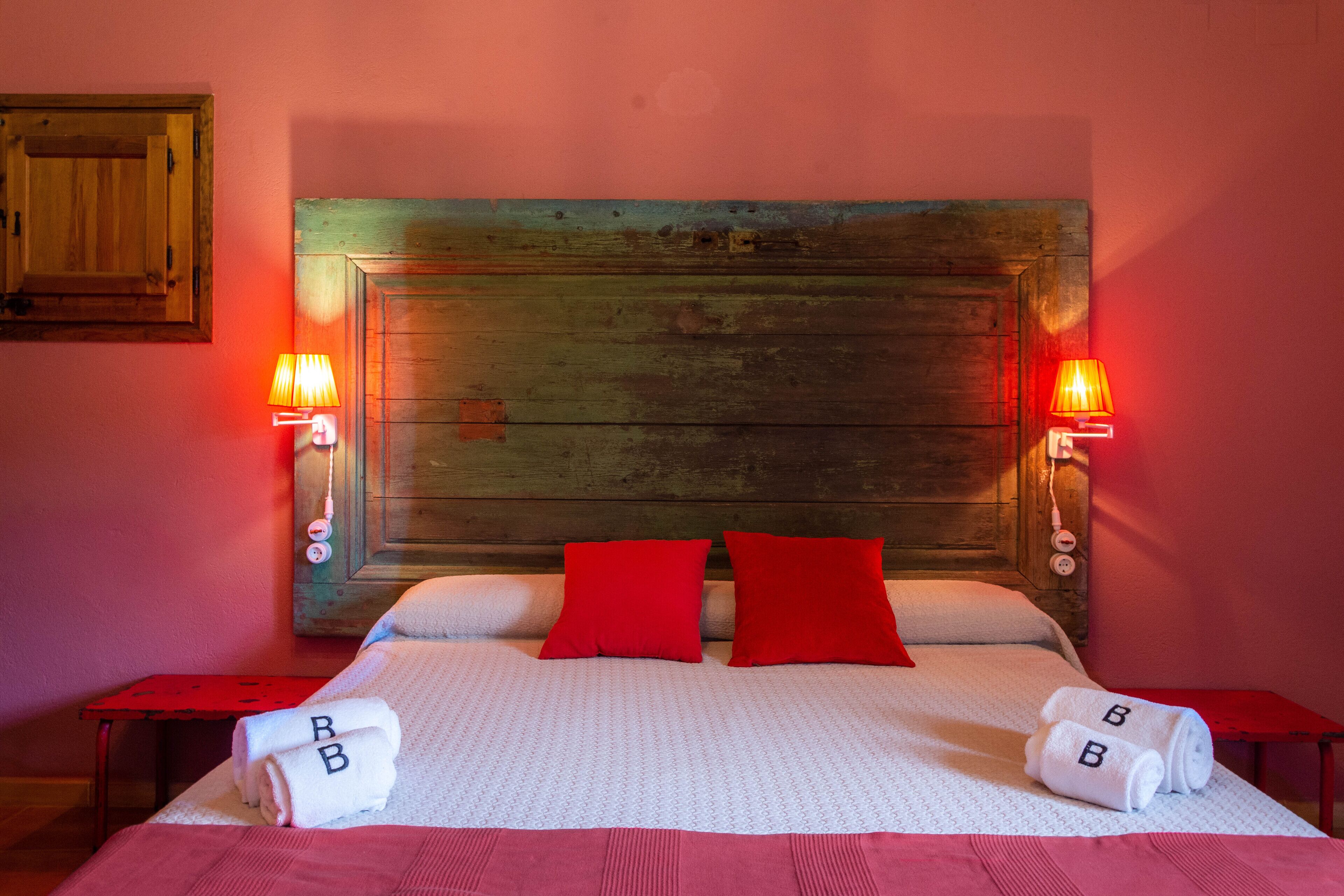 A hotel room with a wooden headboard, two red pillows, a white bedspread, and two bedside tables with rolled towels.