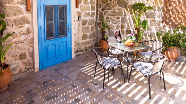 A stone building with a blue door, a wooden ceiling, and a tiled patio with chairs and potted plants. - Galini Apartment (Rhodes)