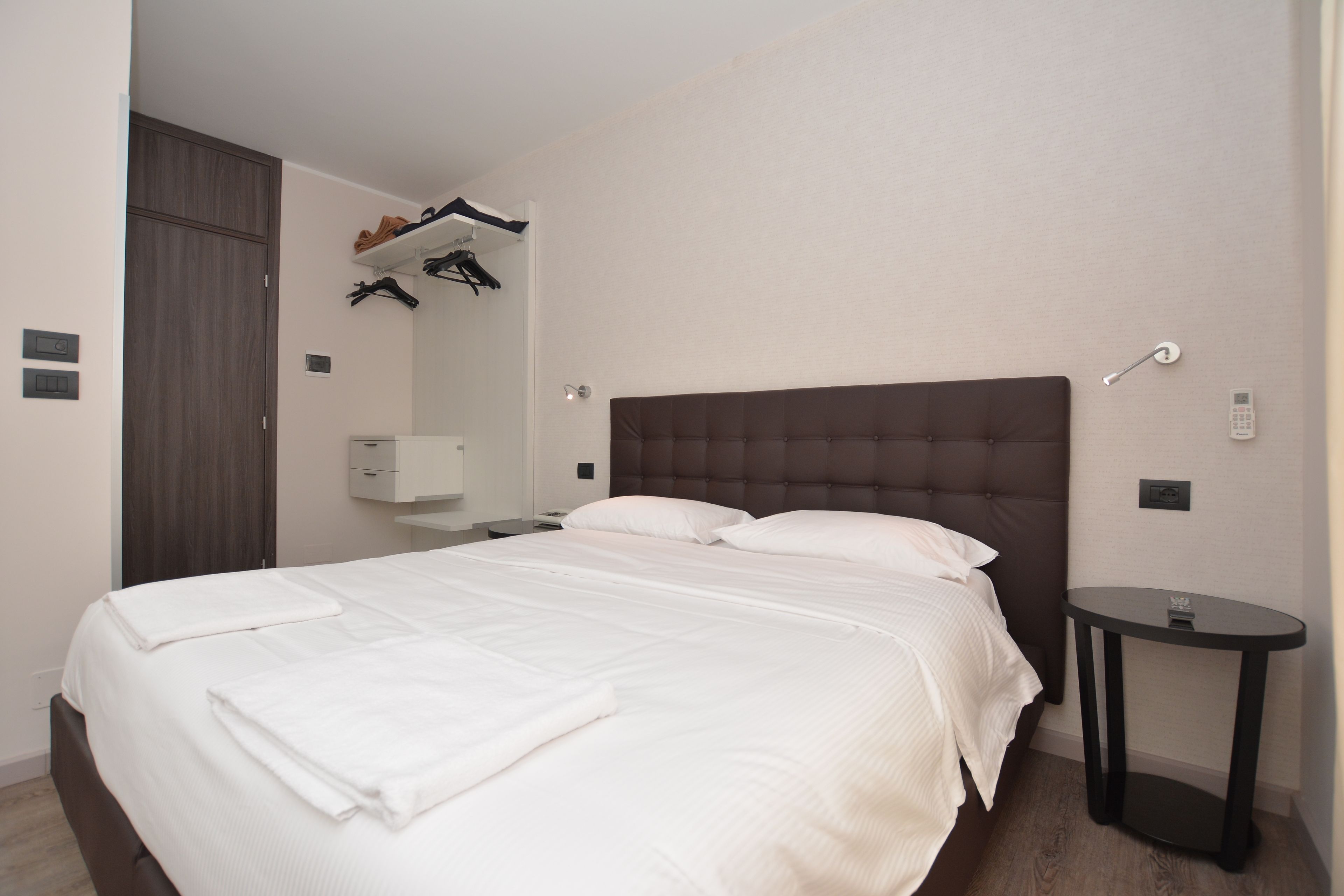 A neatly made bed with white linens and a folded towel, a bedside table with a lamp, and a wardrobe with hanging clothes.
