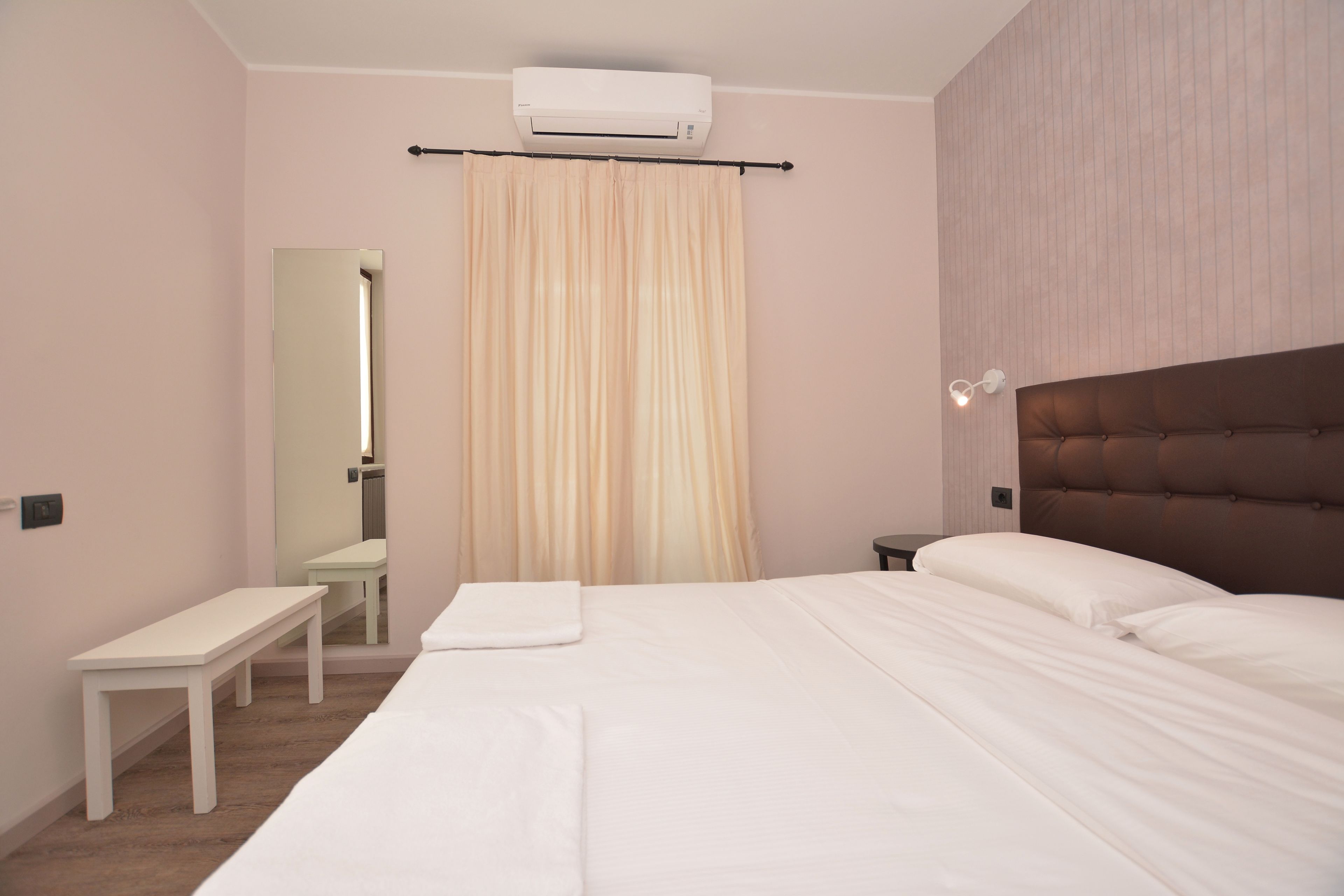 A hotel room with a large bed, a bedside table, a bench, a mirror, and a wall-mounted air conditioner.