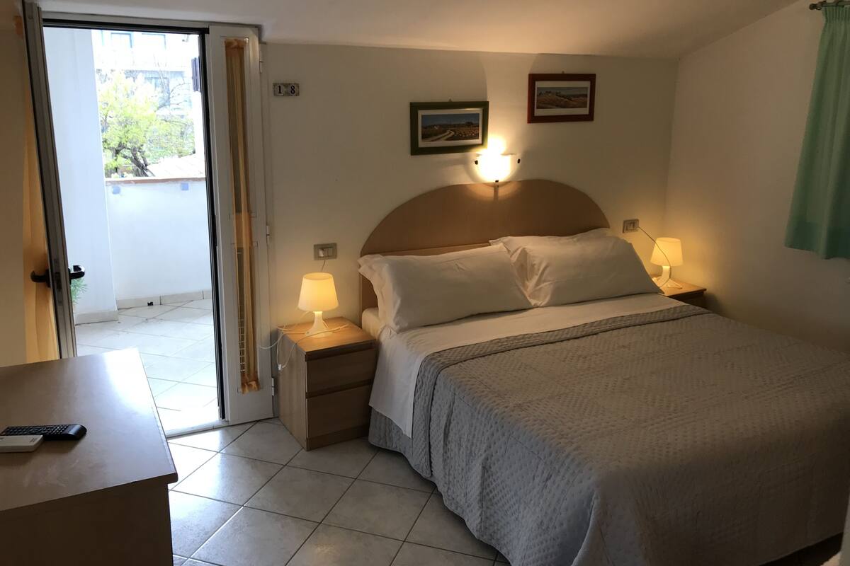 Habitación de hotel con cama, lámparas de noche, mesita de noche, escritorio con teclado y vista al balcón exterior.