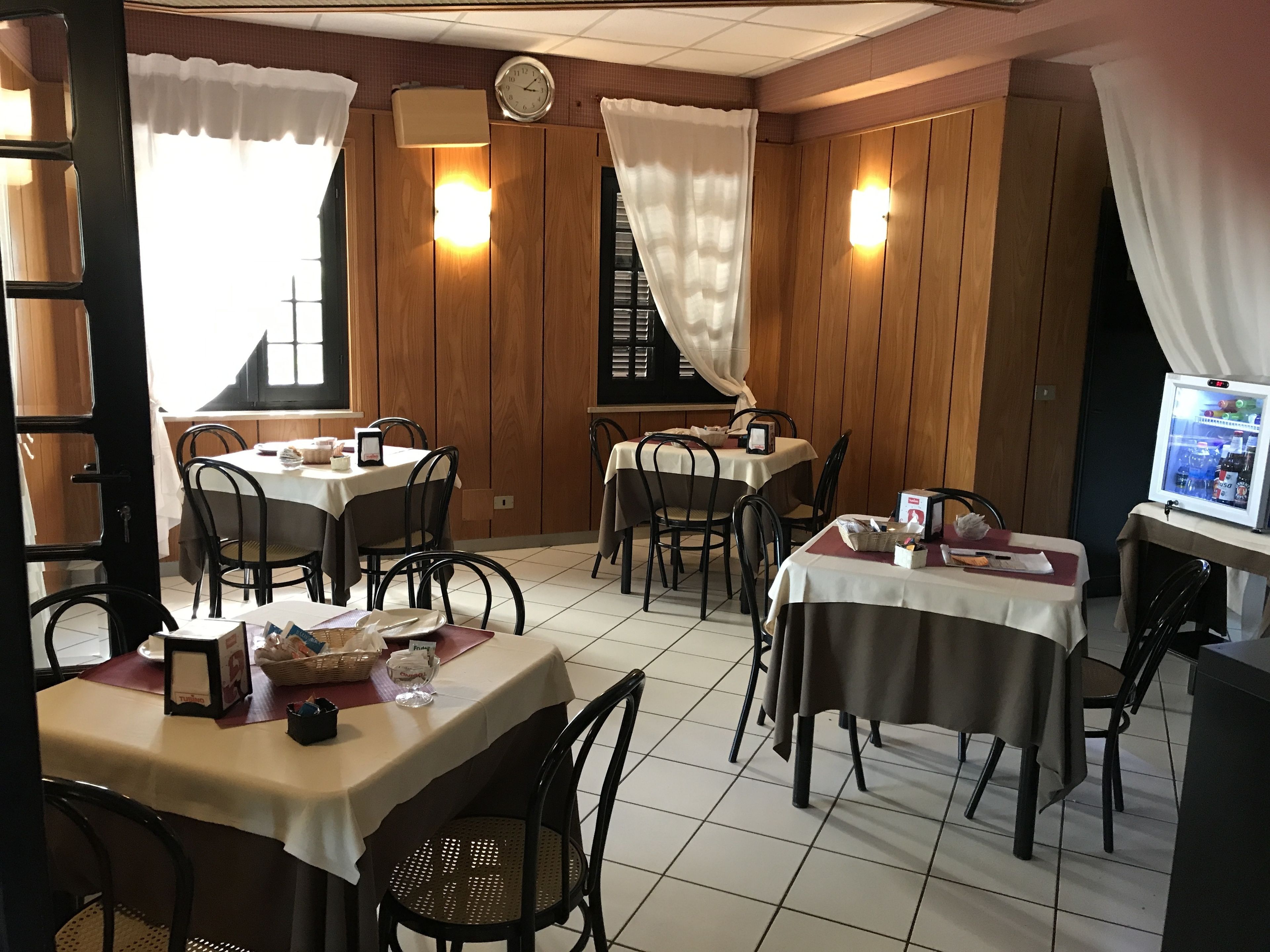 A restaurant with wooden panel walls, multiple tables with white tablecloths, black chairs, and a refrigerator with a digital display.