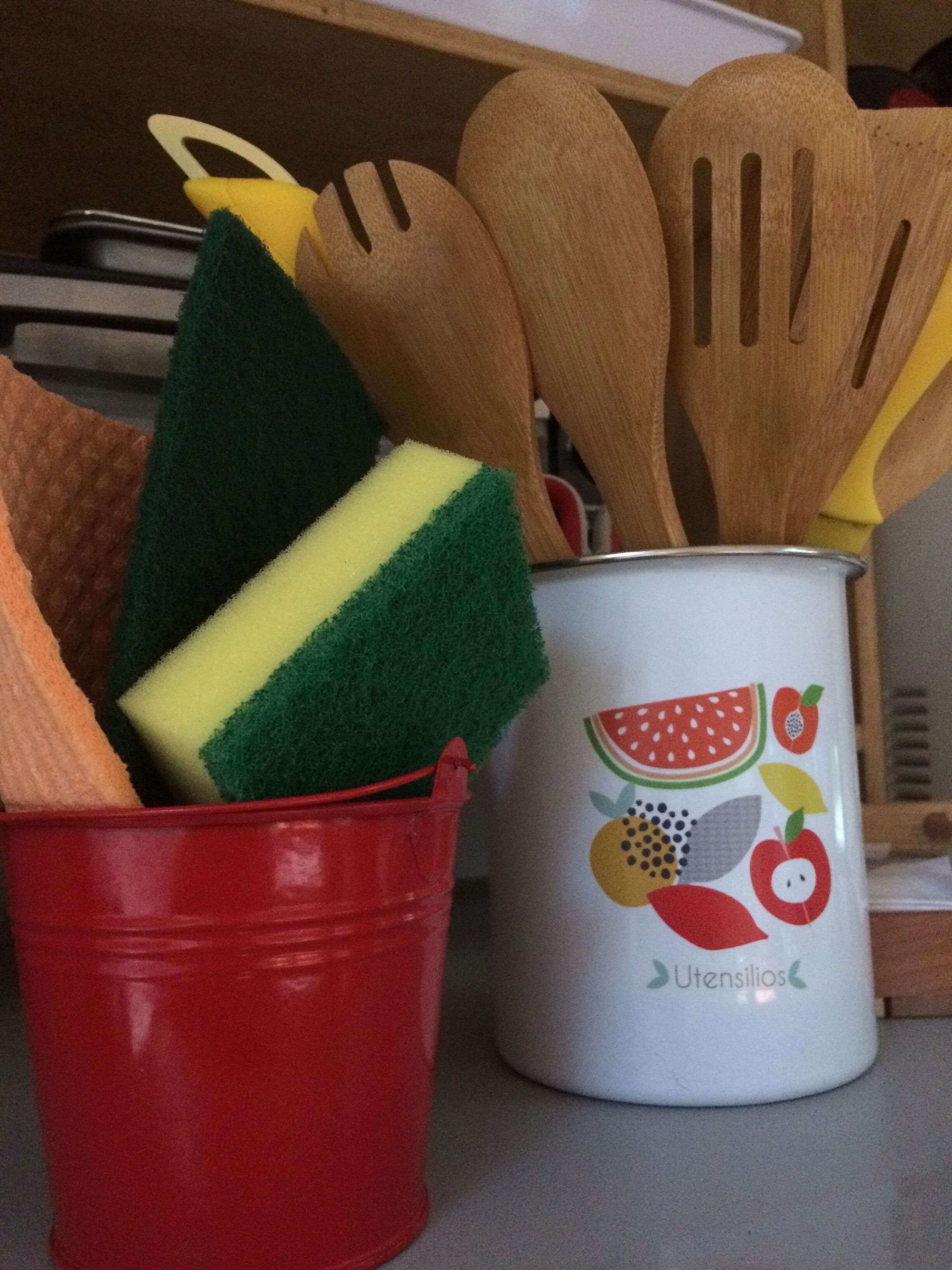A kitchen utensil holder with wooden spatulas and a red bucket containing a green sponge.