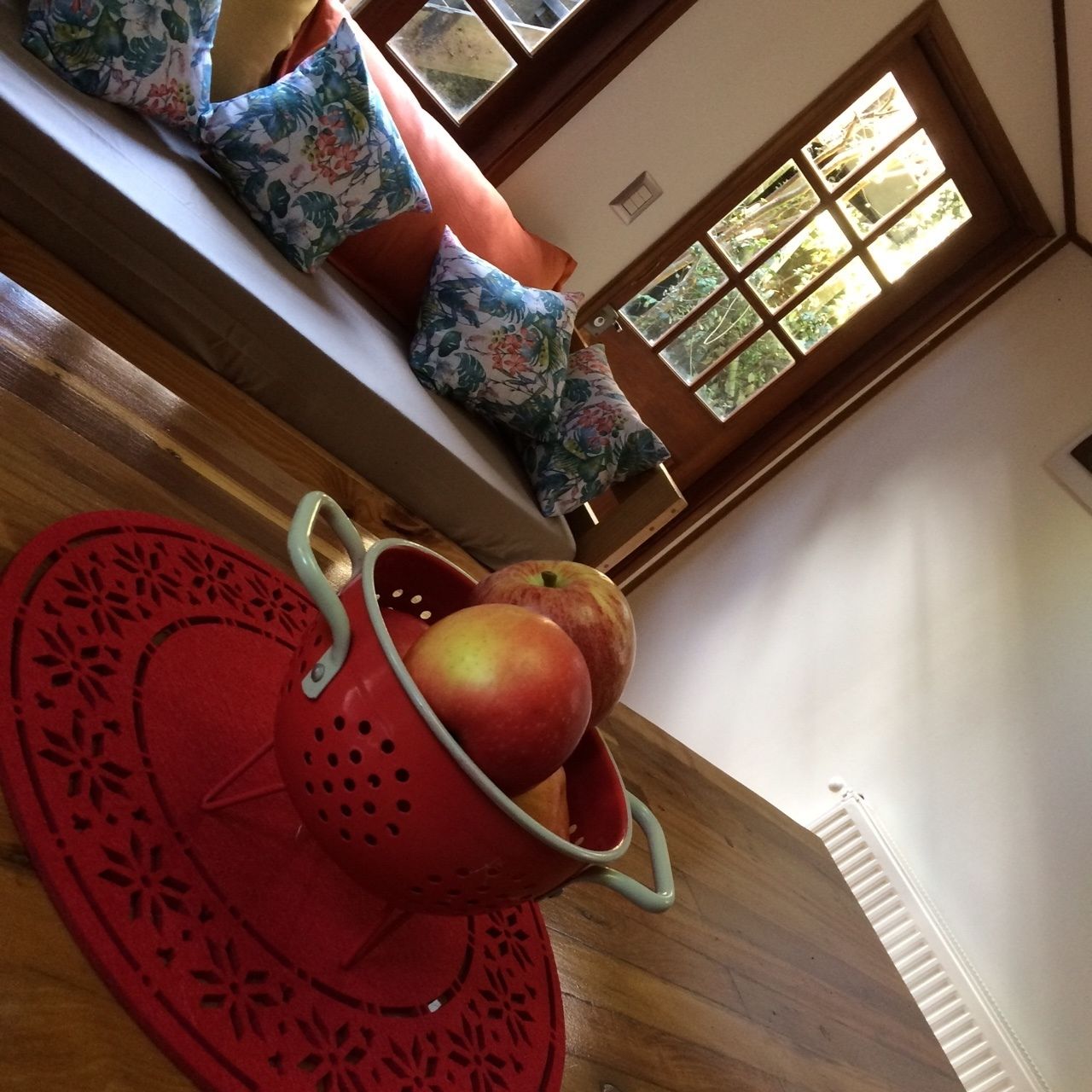 Une pièce avec un plancher en bois, un tapis rouge, un panier de fruits contenant des pommes et des coussins à motifs floraux sur un lit.