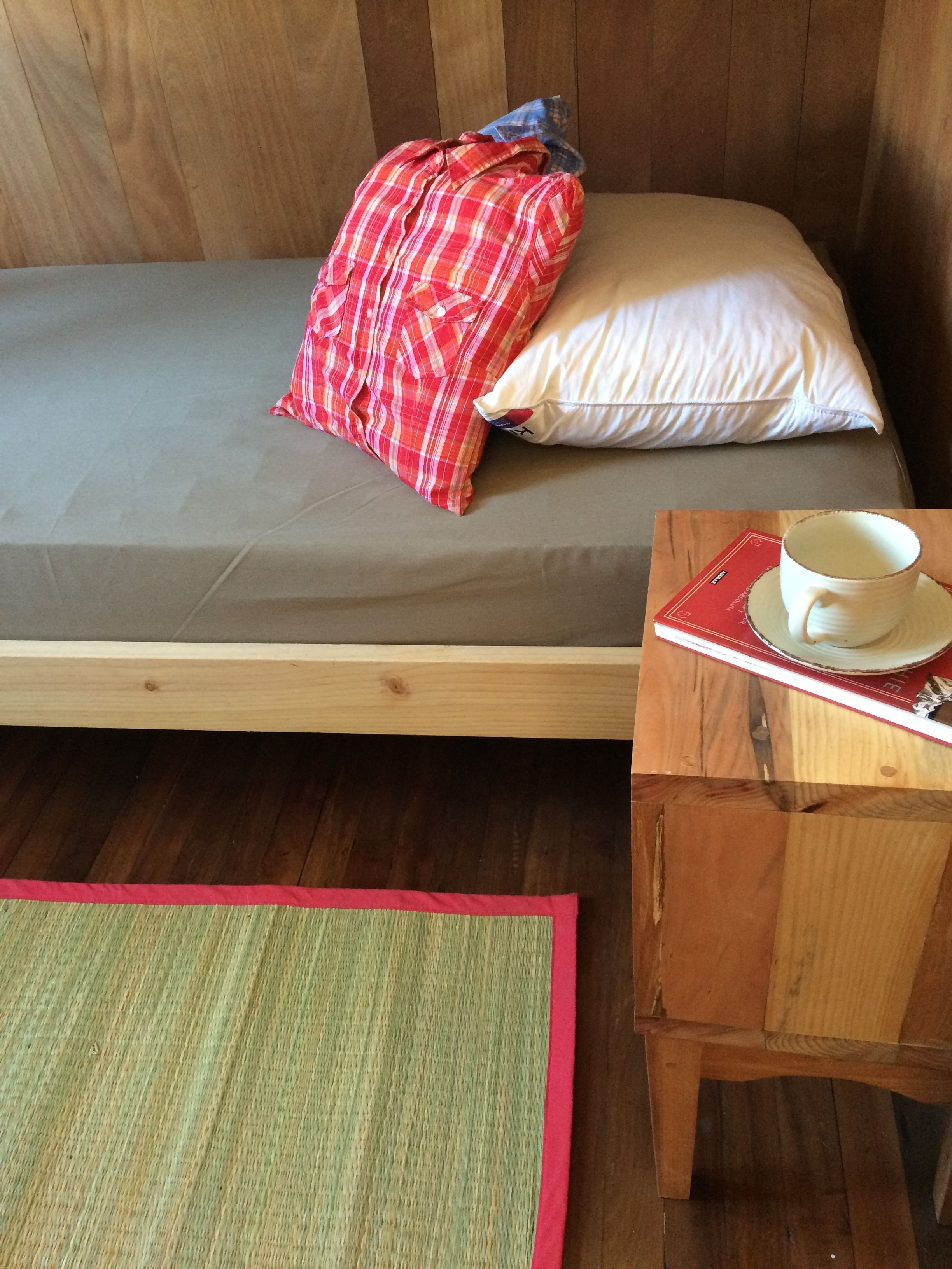 Una cama individual con colchón gris, un cojín de cuadros rojos y uno blanco. Hay una mesita de noche de madera con una taza y un platillo, y un libro rojo. Un tapete verde y rojo está frente a la cama.