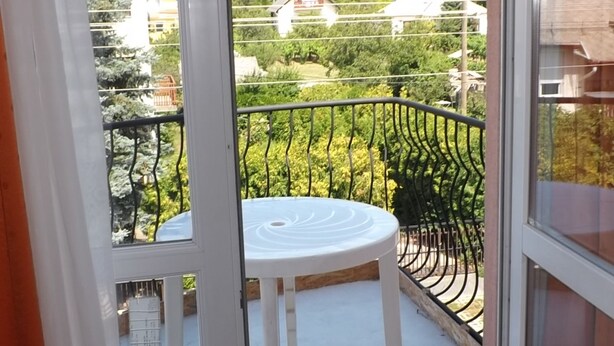 En balkon med hvidt bord og stole, gelænder i metal og udsigt over et boligområde med huse og grønt.
