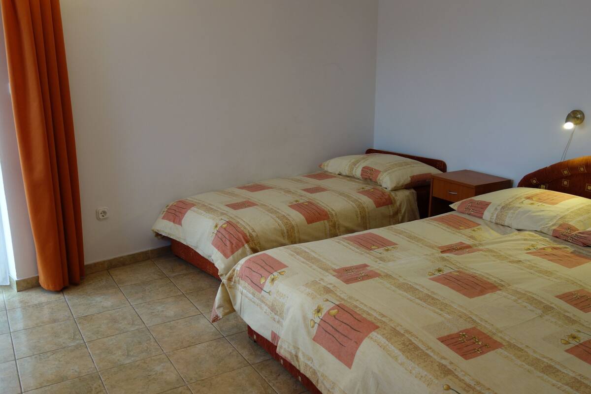 A bedroom with two beds, a nightstand, a wall socket, and a window with curtains.