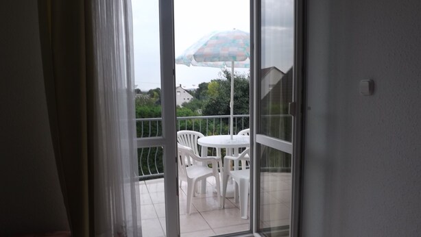 Een balkon met een witte tafel en stoelen, een glazen deur en uitzicht op groen en gebouwen.