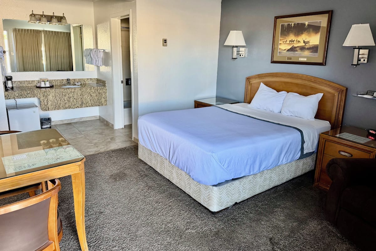 Una habitación de hotel con cama, mesitas de noche, escritorio, silla y baño con lavamanos, espejo y toallero.