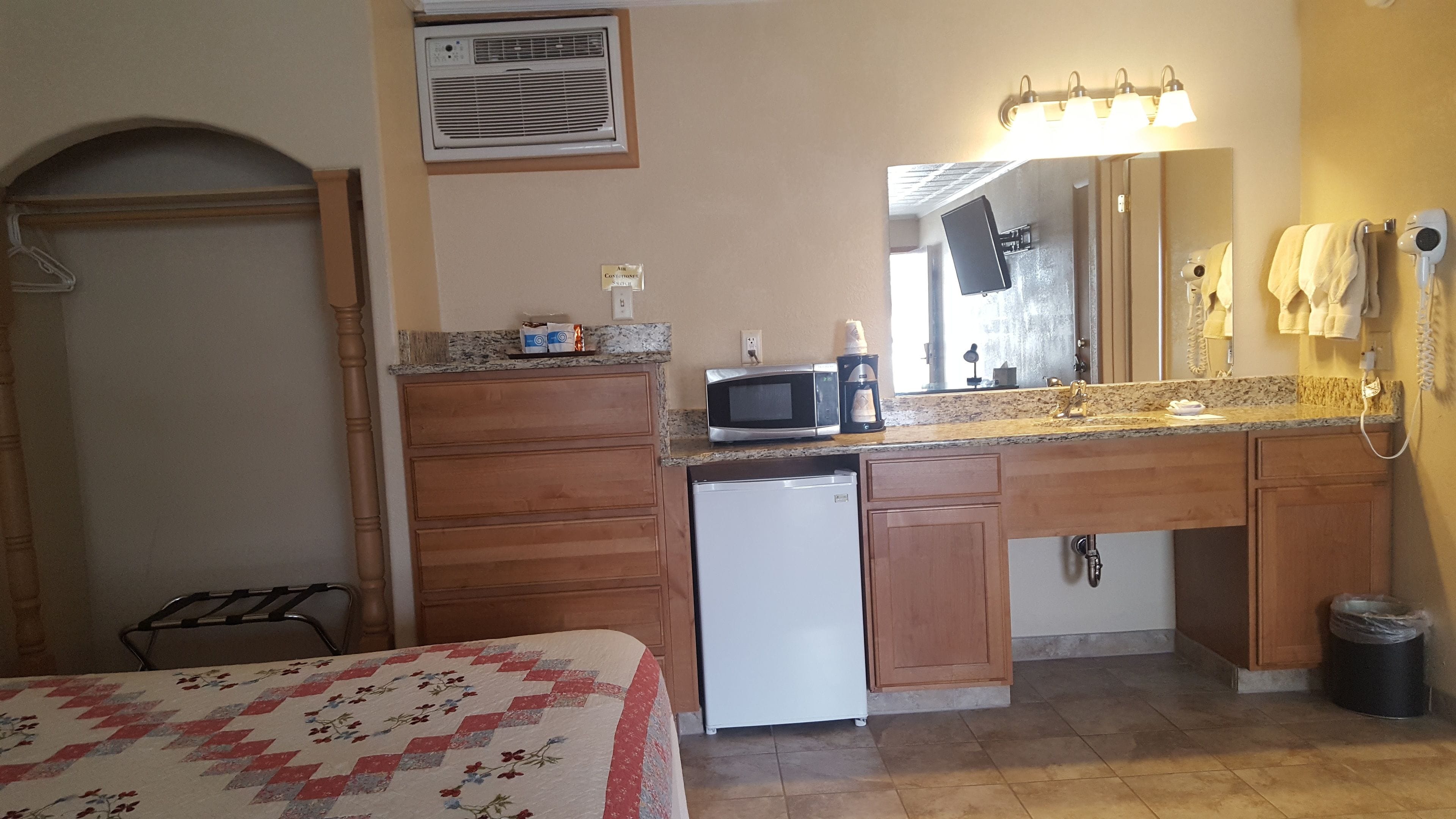 Habitación de hotel pequeña con kitchenette, una cama con colcha estampada, y un baño con lavamanos, espejo y perchero.