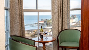 A hotel room with a bed, two chairs, a small table with a bottle and glass, and a view of the sea and buildings. - Royal Lion Hotel (Tenby)