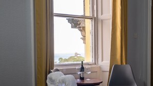 A hotel room with a bed, a table with a bottle and glass, a chair, and a window with curtains. - Royal Lion Hotel (Tenby)