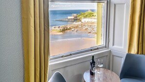 A room with a large window offering a view of a beach and pier, a small round wooden table with a bottle and glasses, and two modern chairs. - Royal Lion Hotel (Tenby)