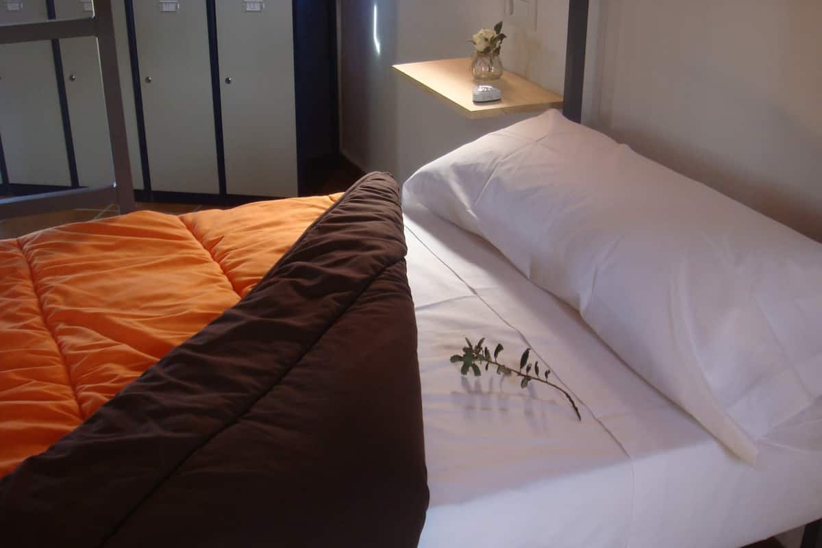 A bed with an orange and brown quilt and white sheets, a small bedside table with a vase of flowers, and a wardrobe in the background.