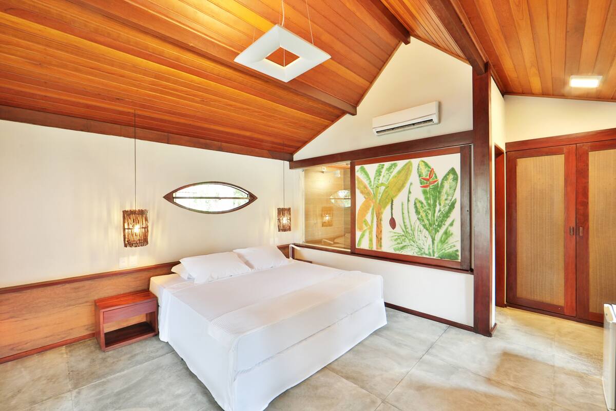 A bedroom with a large bed, wooden ceiling, and a window with a plant painting.