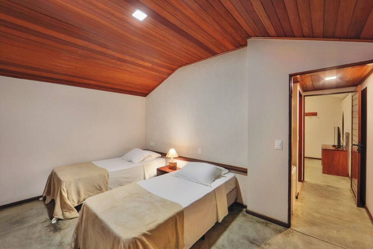 A room with two single beds, a wooden ceiling, a small bedside table with a lamp, and a door leading to another room.