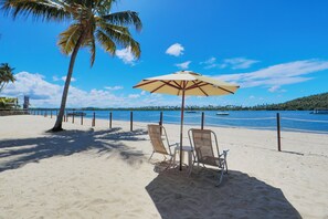 Praia com duas cadeiras de praia e um guarda-sol grande sob uma palmeira, com vista para um mar calmo e colinas ao longe.