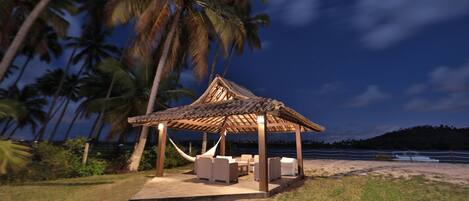 A wooden gazebo with a thatched roof, surrounded by palm trees, and overlooking a beach.