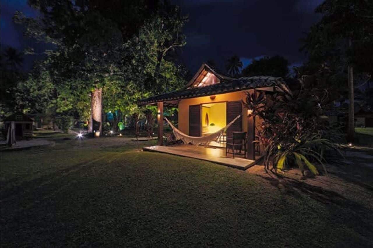 Een klein, gezellig huis met een veranda en een hangmat, omringd door bomen en een goed verlichte tuin.