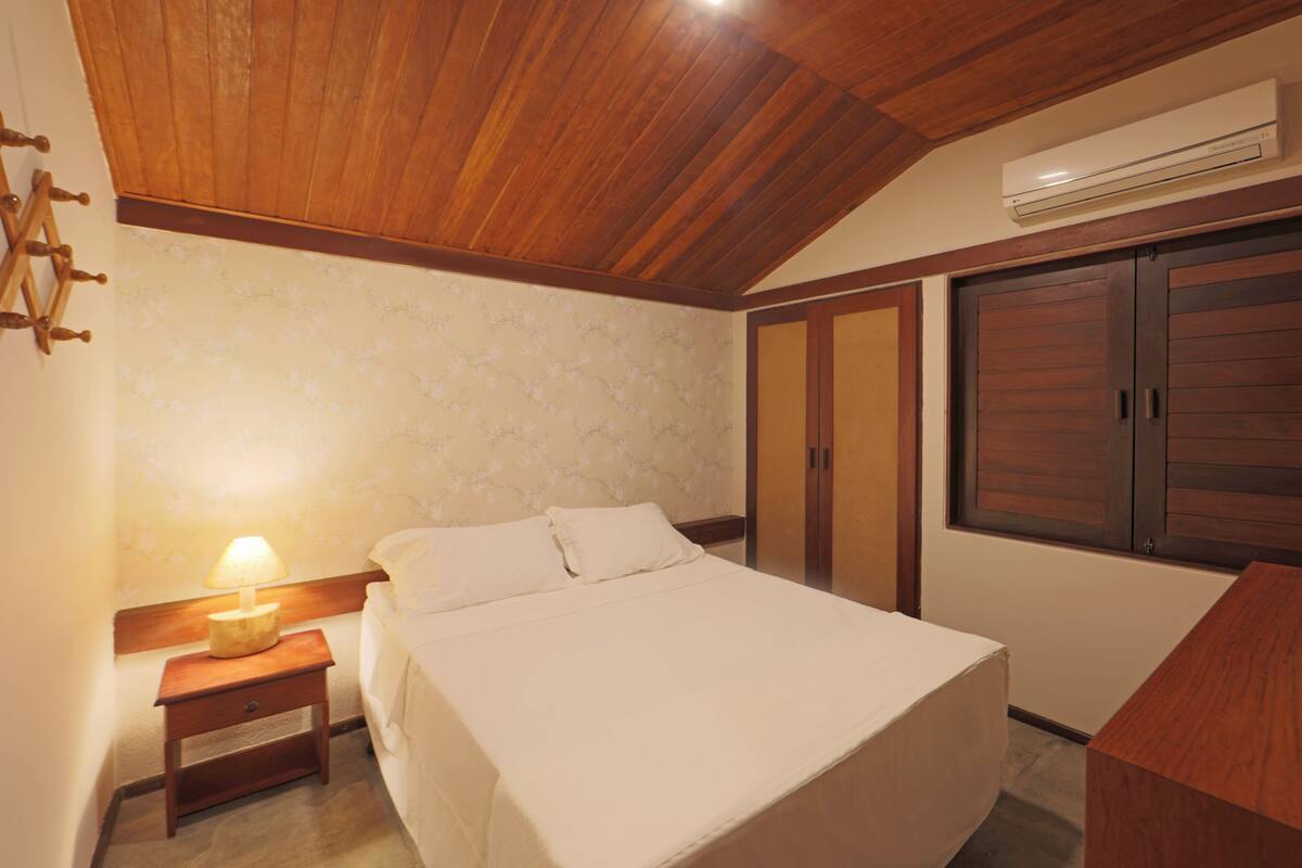 A bedroom with a wooden ceiling, a bed with white linens, a nightstand with a lamp, and a wardrobe.