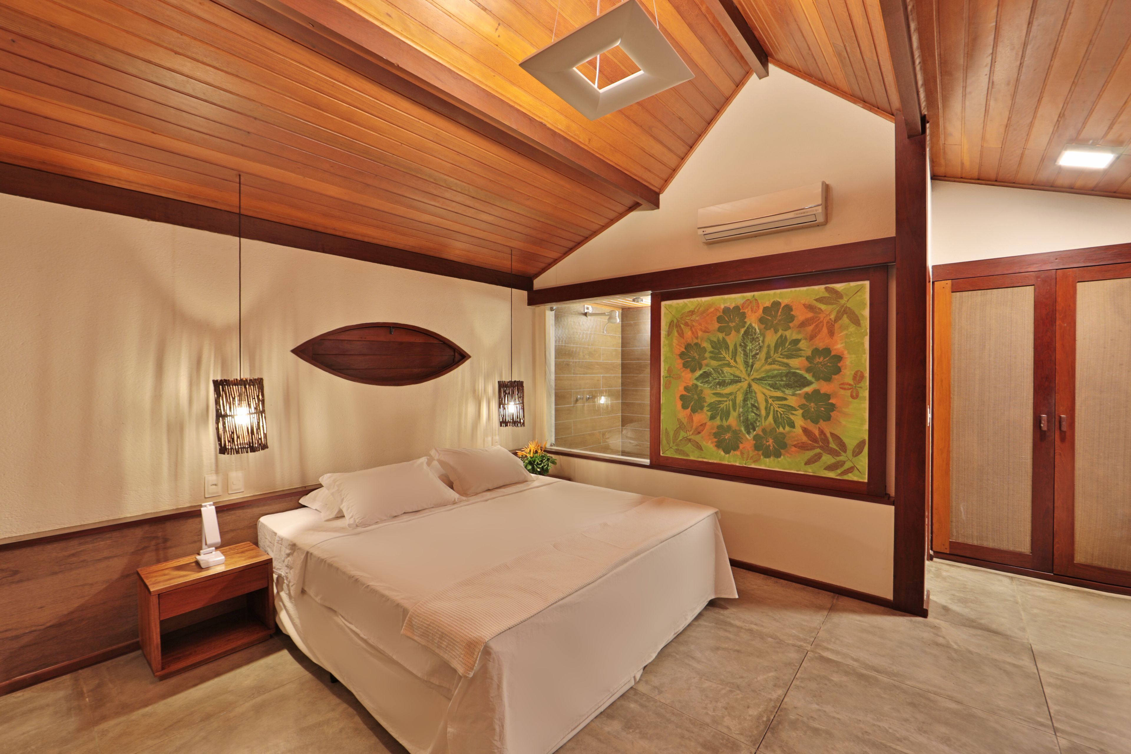 A bedroom with a wooden ceiling, a large bed, a nightstand, a wall-mounted lamp, and a large floral painting.