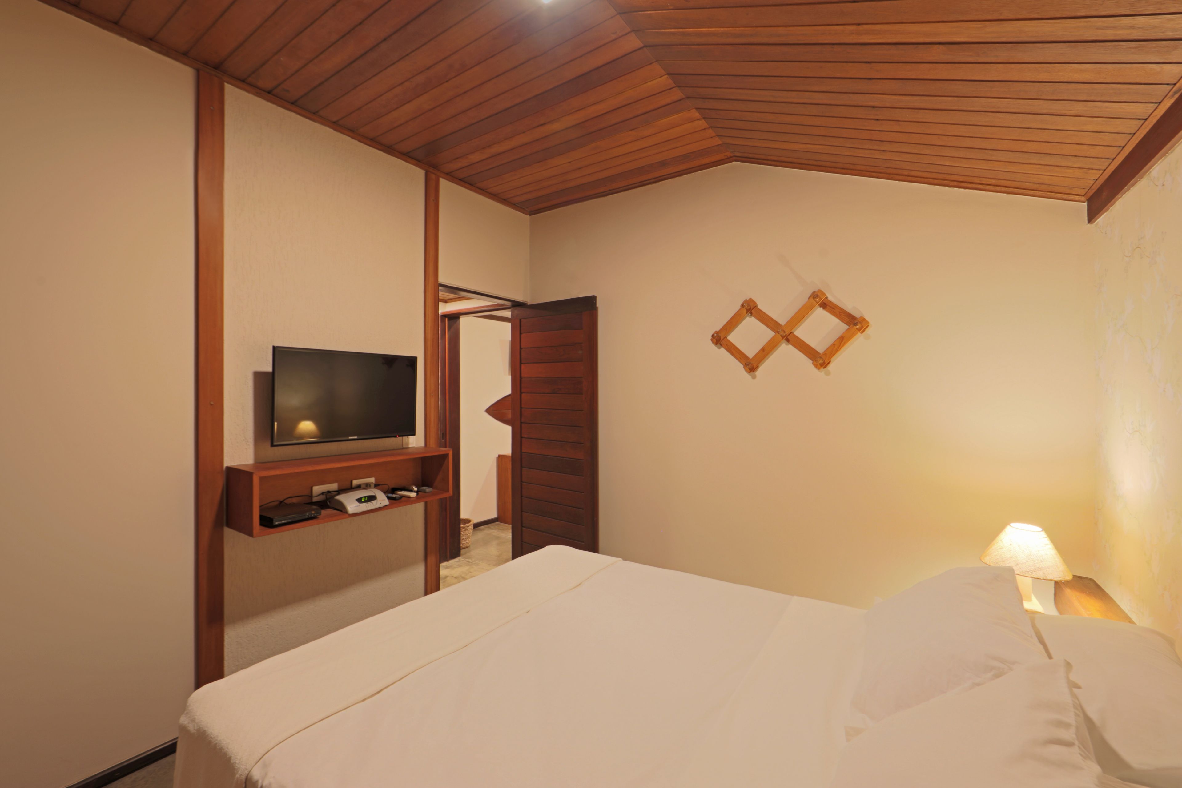 A hotel room with a bed, a TV mounted on a wooden shelf, a bedside lamp, and a wooden ceiling.