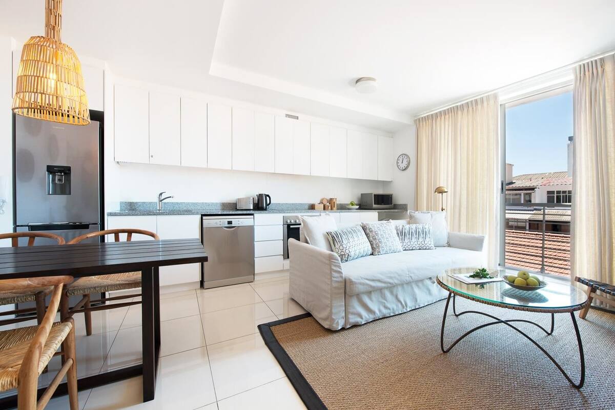 Une cuisine moderne avec des meubles blancs, un coin repas avec une table et des chaises en bois, un canapé et un petit balcon avec vue.