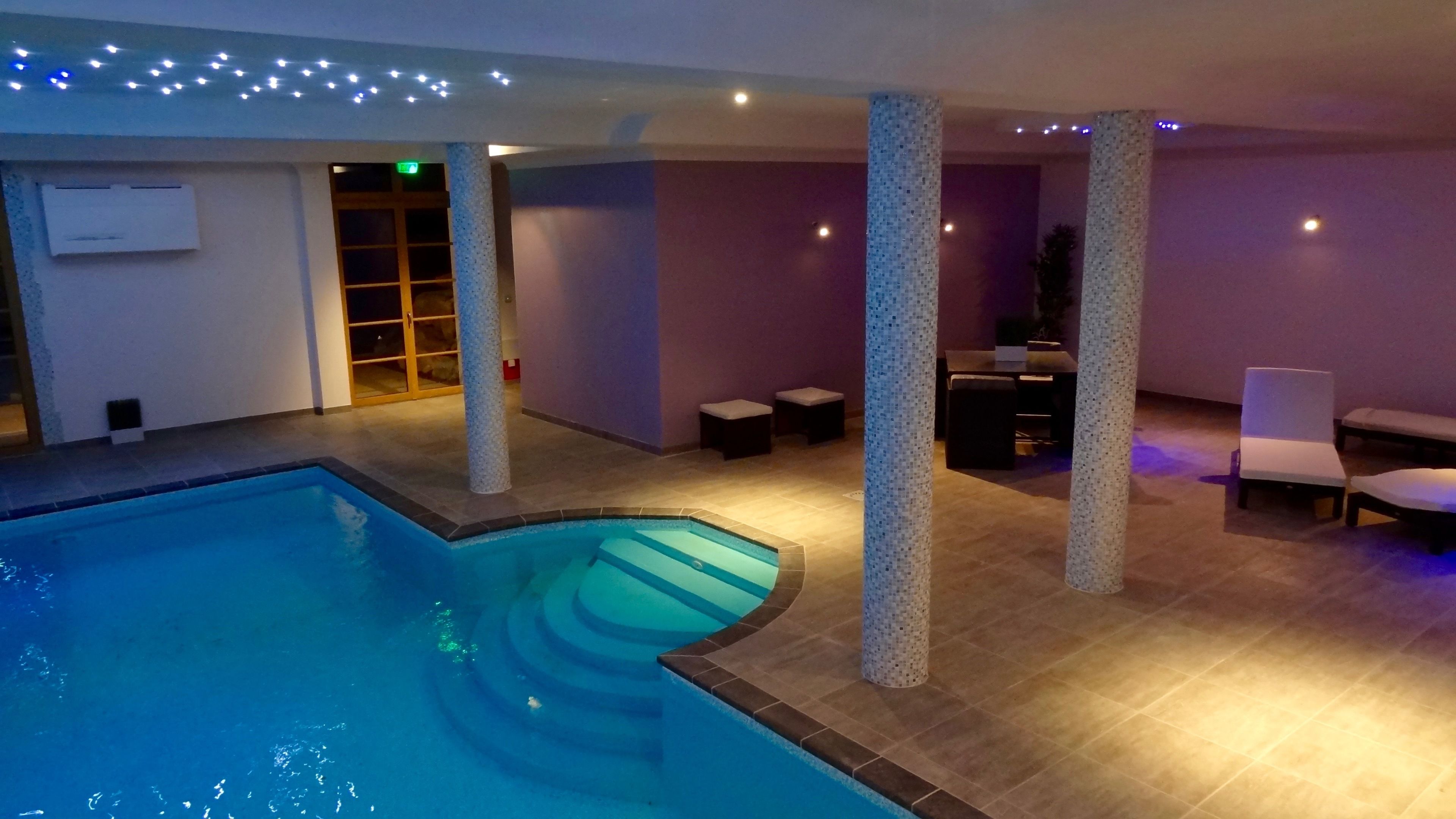 An indoor swimming pool with a tiled floor, a glass door entrance, and a seating area with chairs and a table.