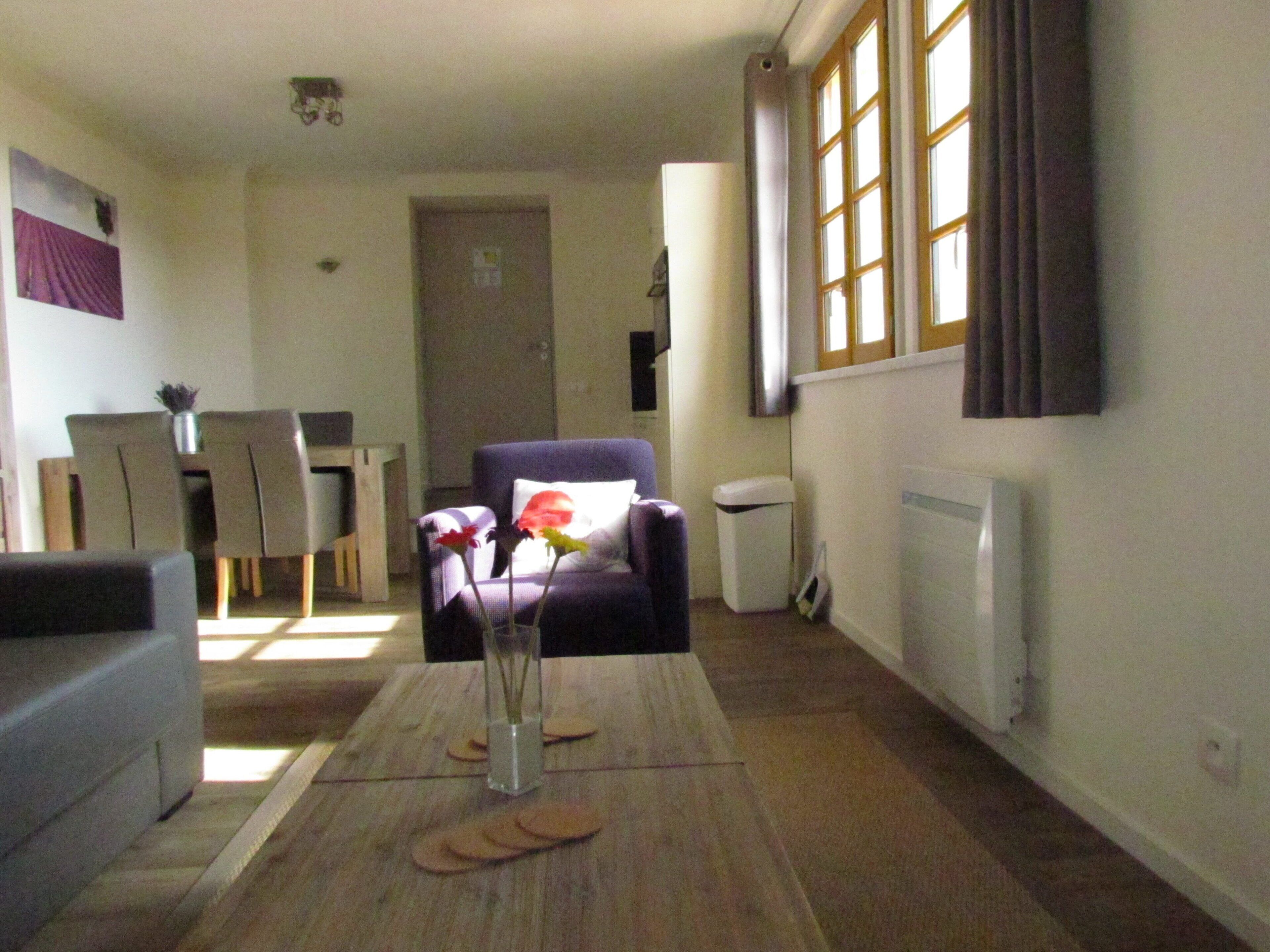 A living room with a sofa, a coffee table with a vase and flowers, a dining area with chairs and a table, and a window with curtains.