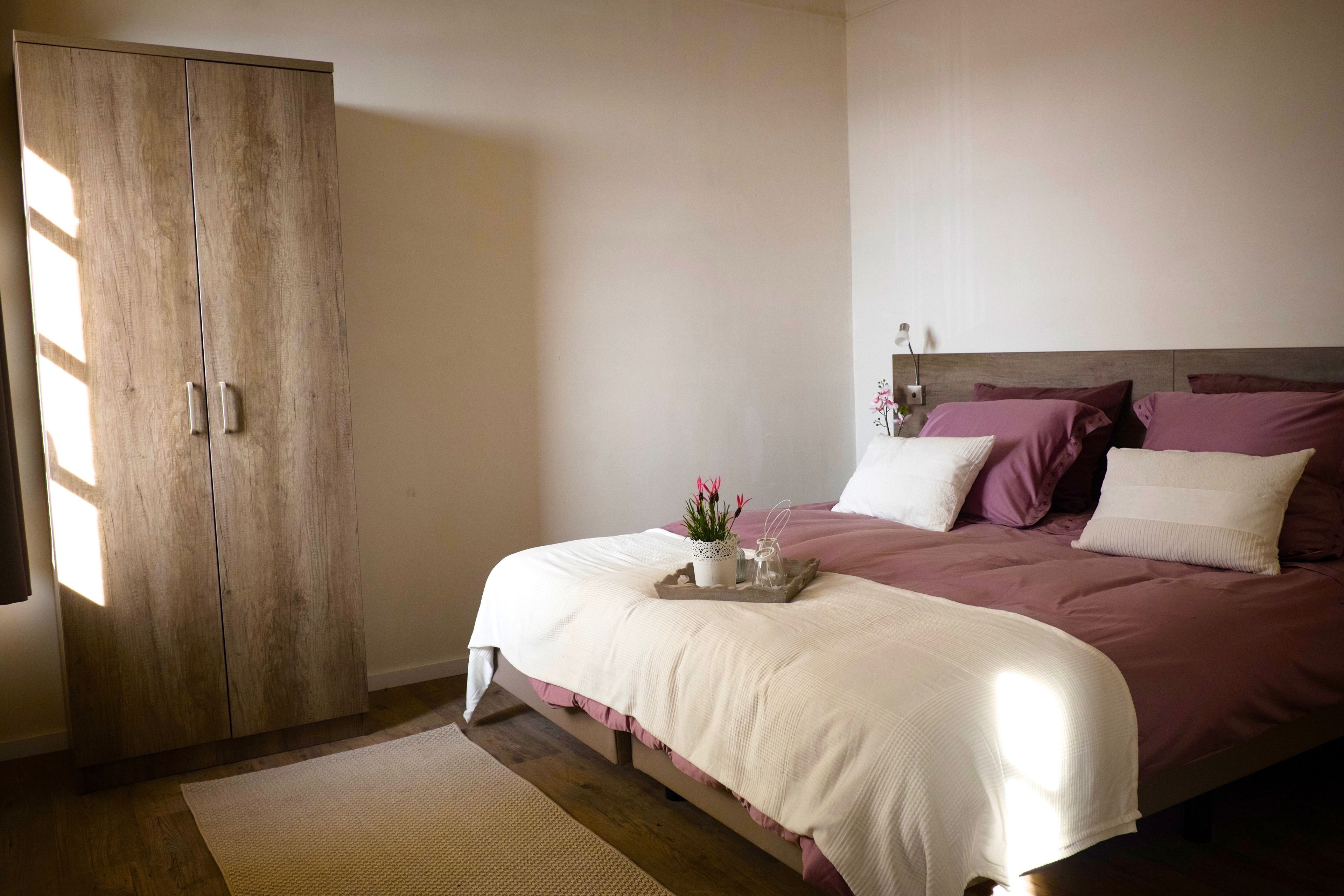 A bedroom with a large wardrobe, a bed with a white and purple bedspread, and a small table with a plant.