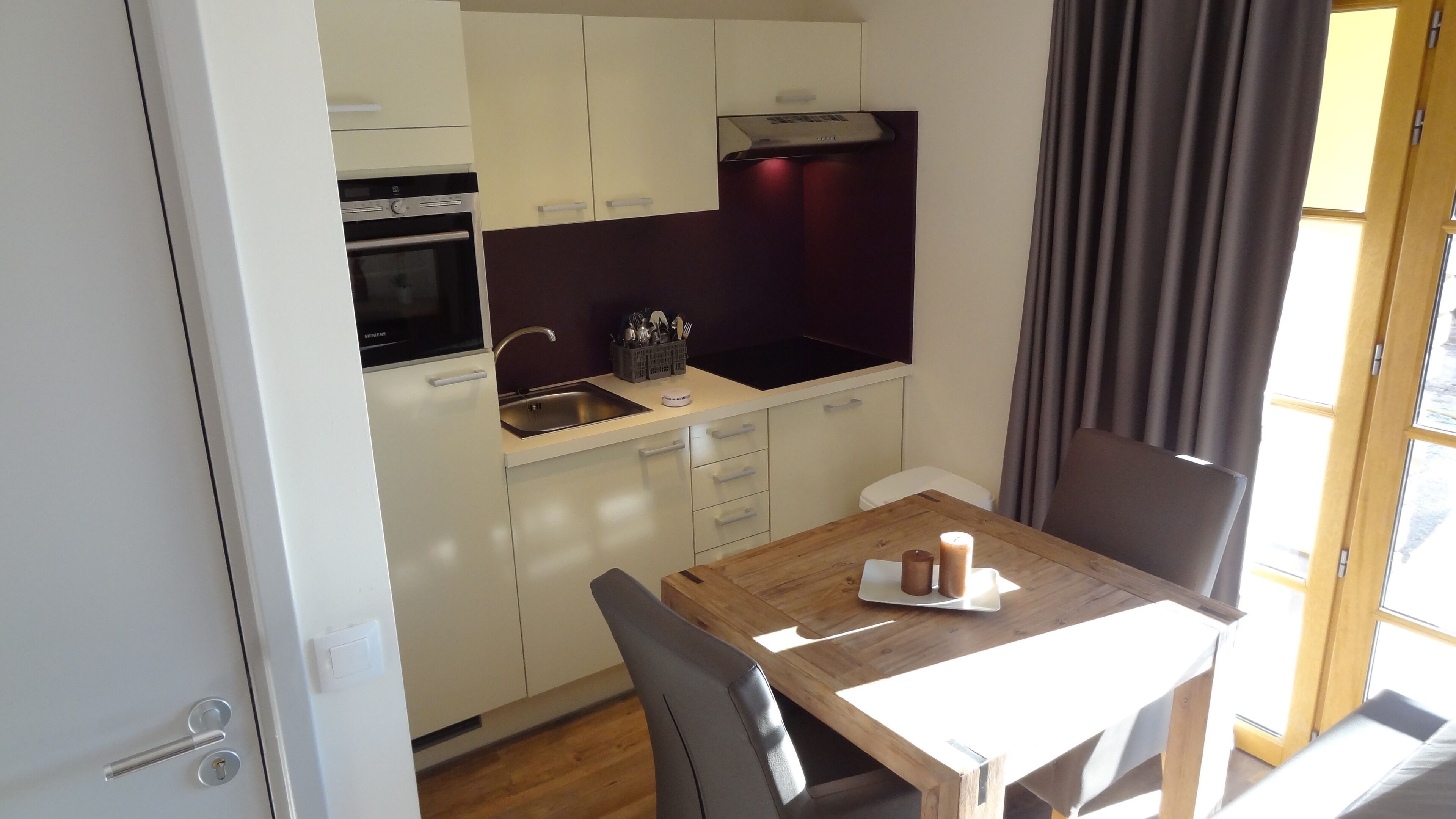 A compact kitchen with white cabinets, a built-in oven, and a sink. There's a wooden table with a candle and a tray, and a chair.