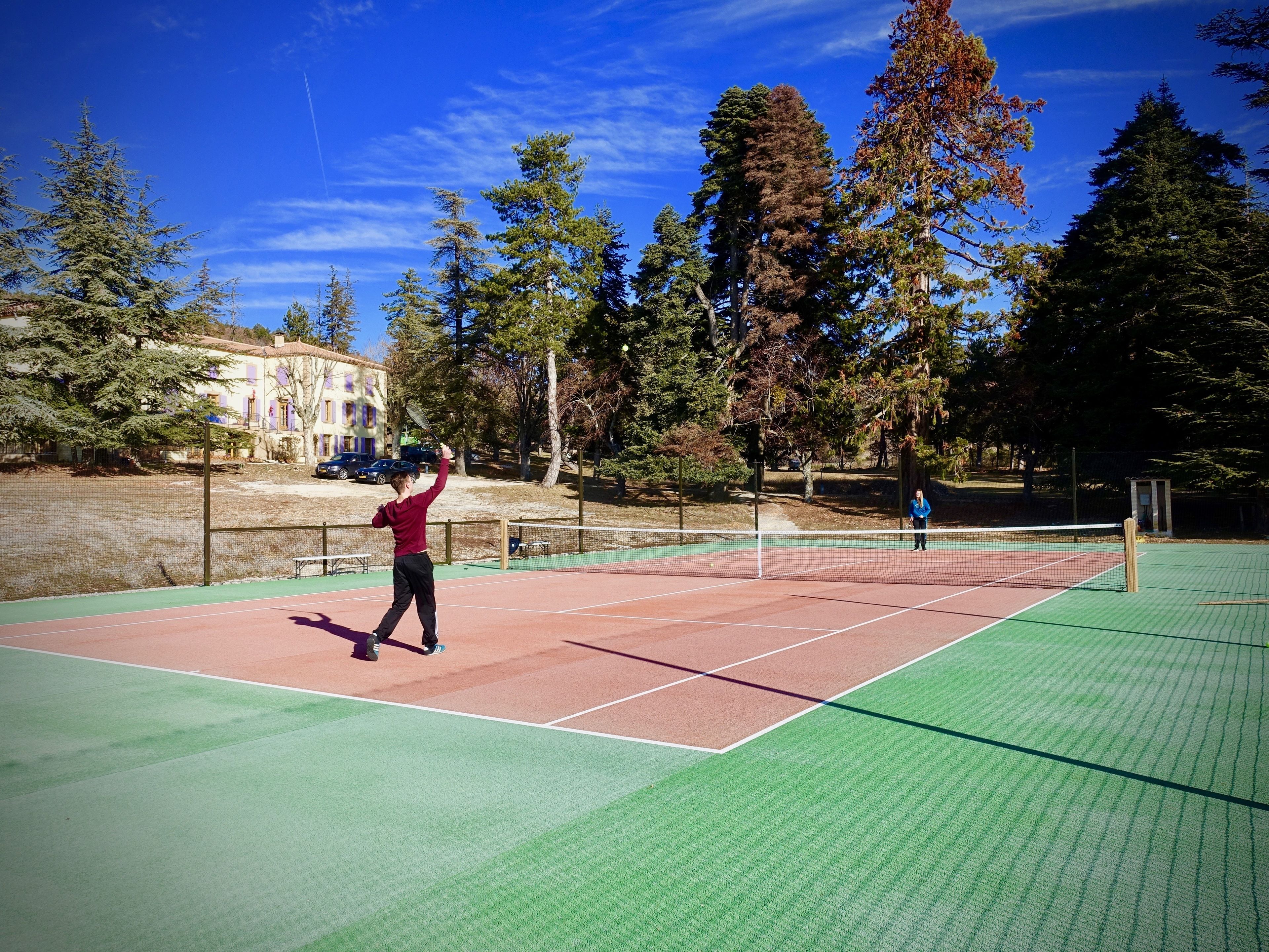 An outdoor tennis court with a net, surrounded by trees and a building in the background.