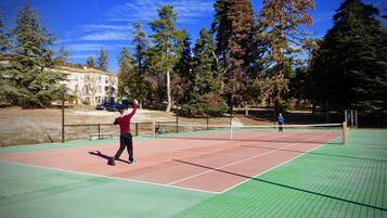 An outdoor tennis court with a net, surrounded by trees and a building in the background.