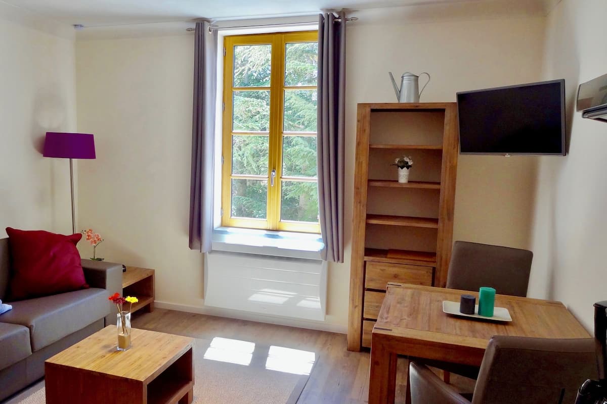Una sala de estar con un sofá, una mesa de centro, un televisor, una mesa de madera y un ventanal con cortinas.