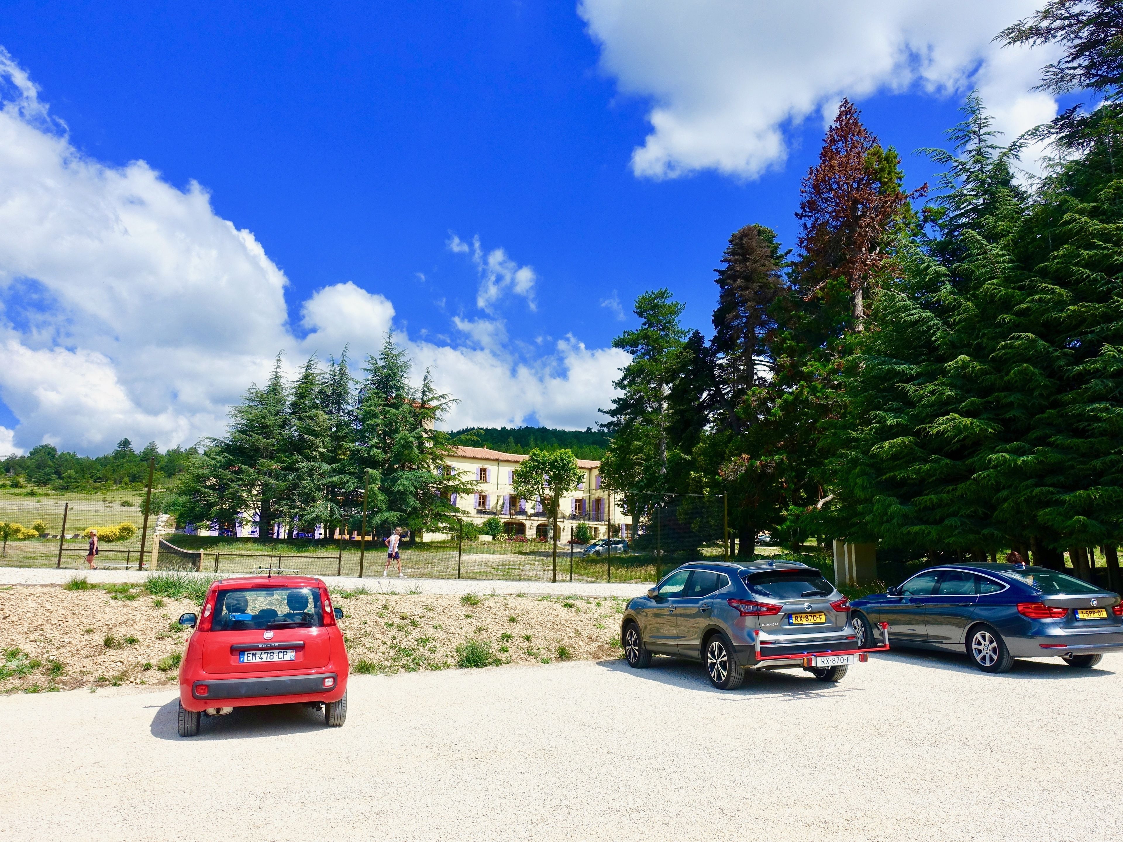 A parking area with three cars, a building with a red roof, and a clear blue sky with clouds.
