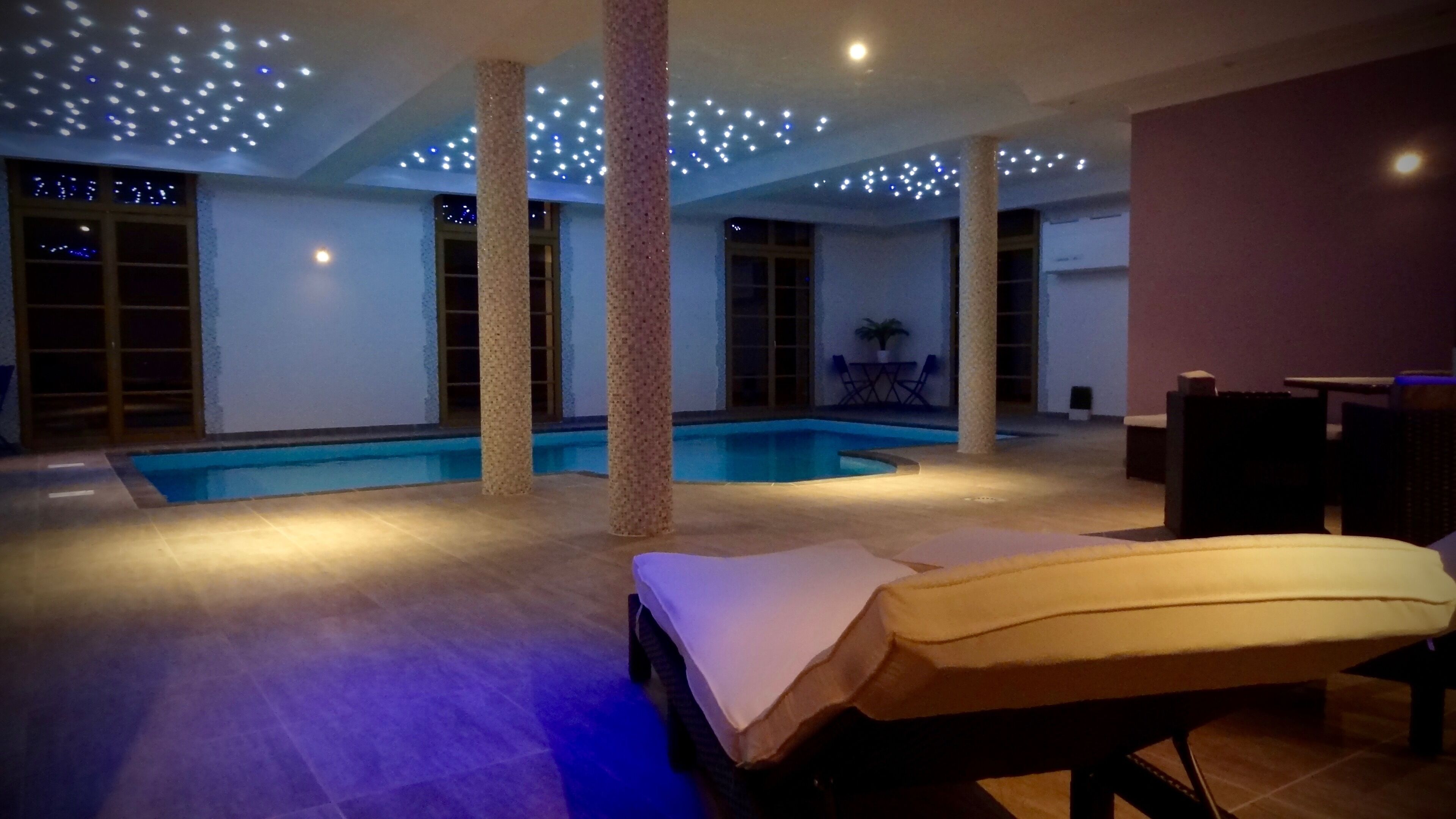 Un espace piscine comprenant des chaises longues, des colonnes et des luminaires au plafond.