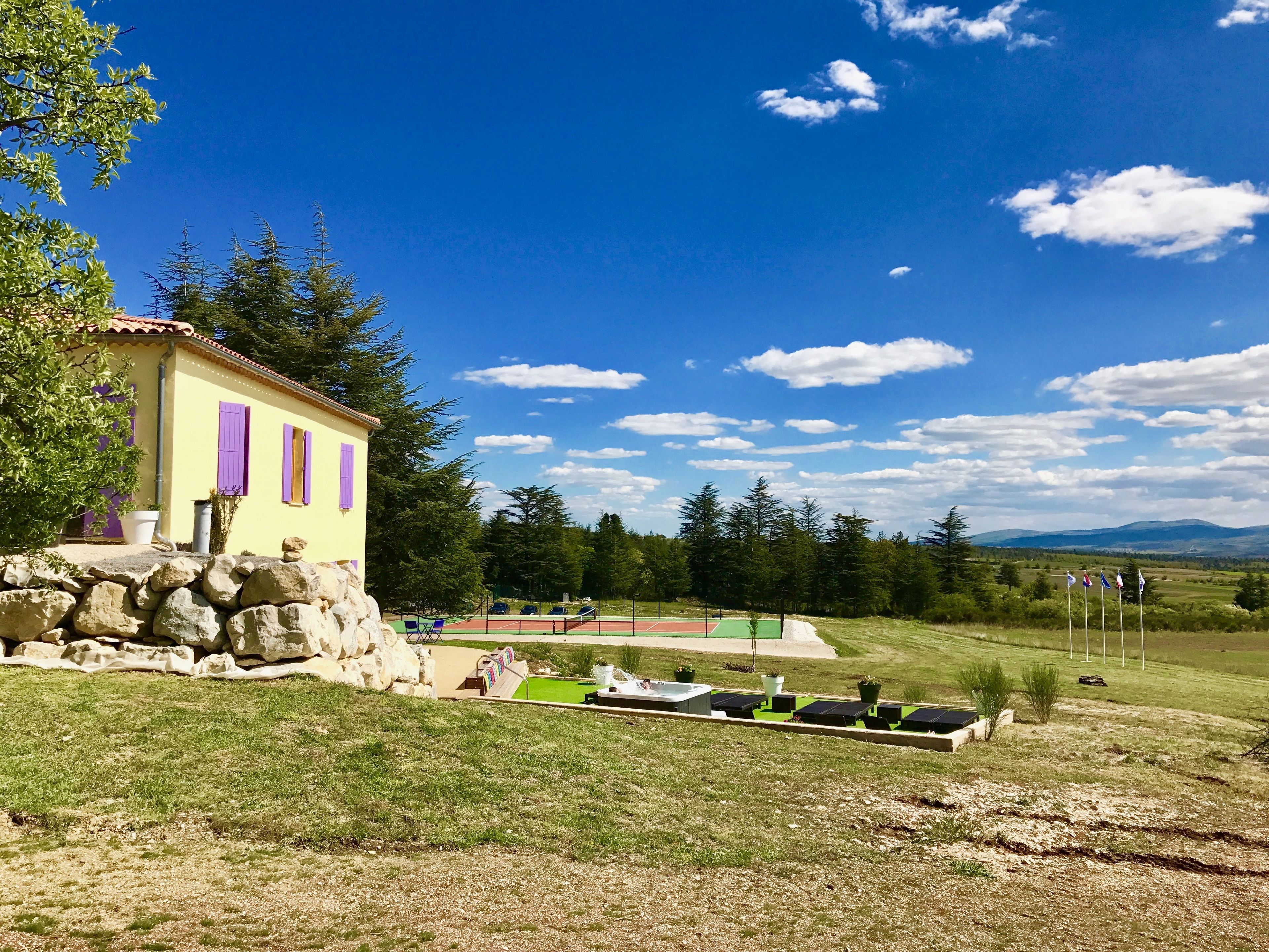 A yellow building with purple shutters, a stone wall, and a tennis court with solar panels.
