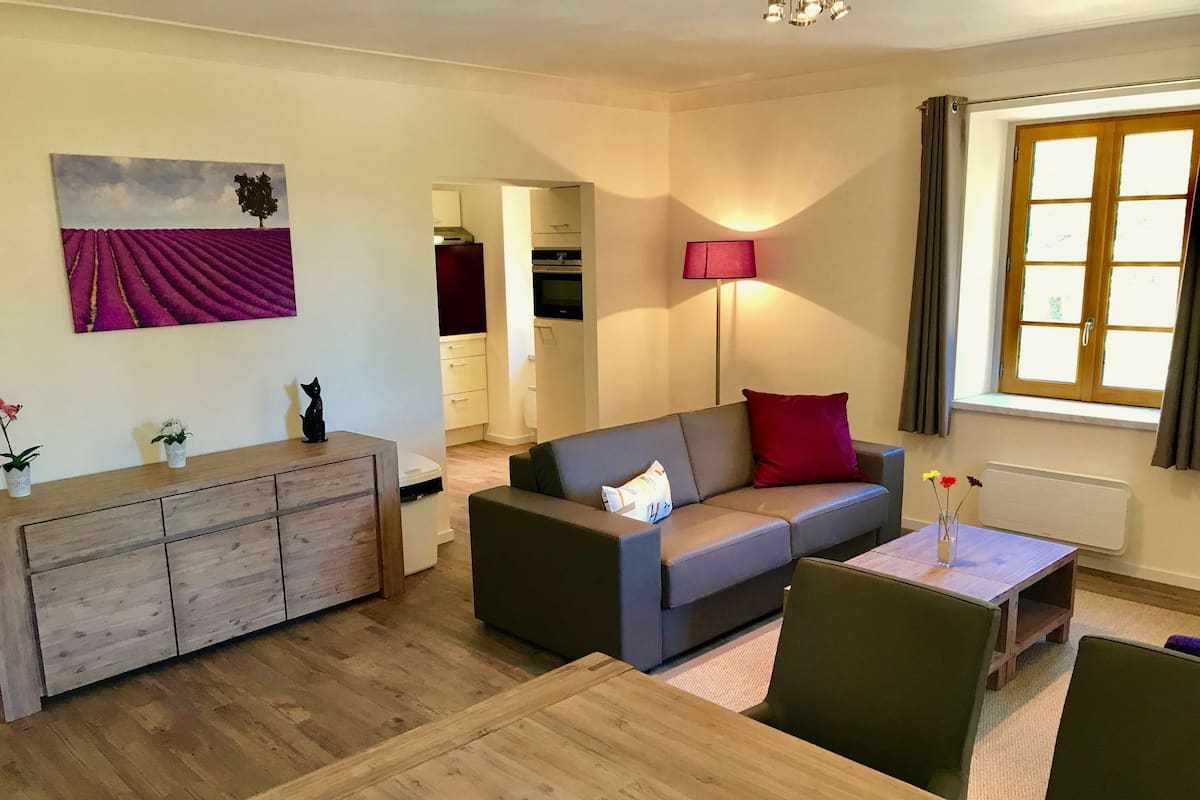 Una sala de estar moderna con un sofá gris, una mesa de comedor de madera y una cocina al fondo.