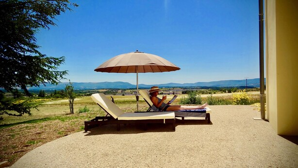A person relaxing on a sun lounger with an umbrella, overlooking a scenic landscape with mountains and fields. - Résidence Château Ferrassières (Ferrassières)