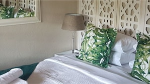 Un lit bien fait, avec une literie blanche et des coussins ornés d’un motif de feuilles vertes, une lampe et un panneau décoratif mural.