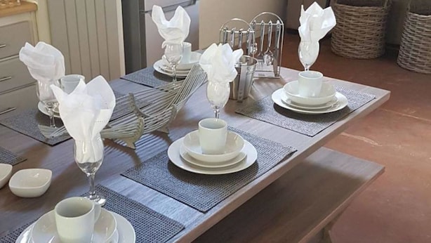 Une table à manger dressée avec de l’assiette blanche, des verres et des serviettes, entourée de paniers en osier et d’un banc en bois.